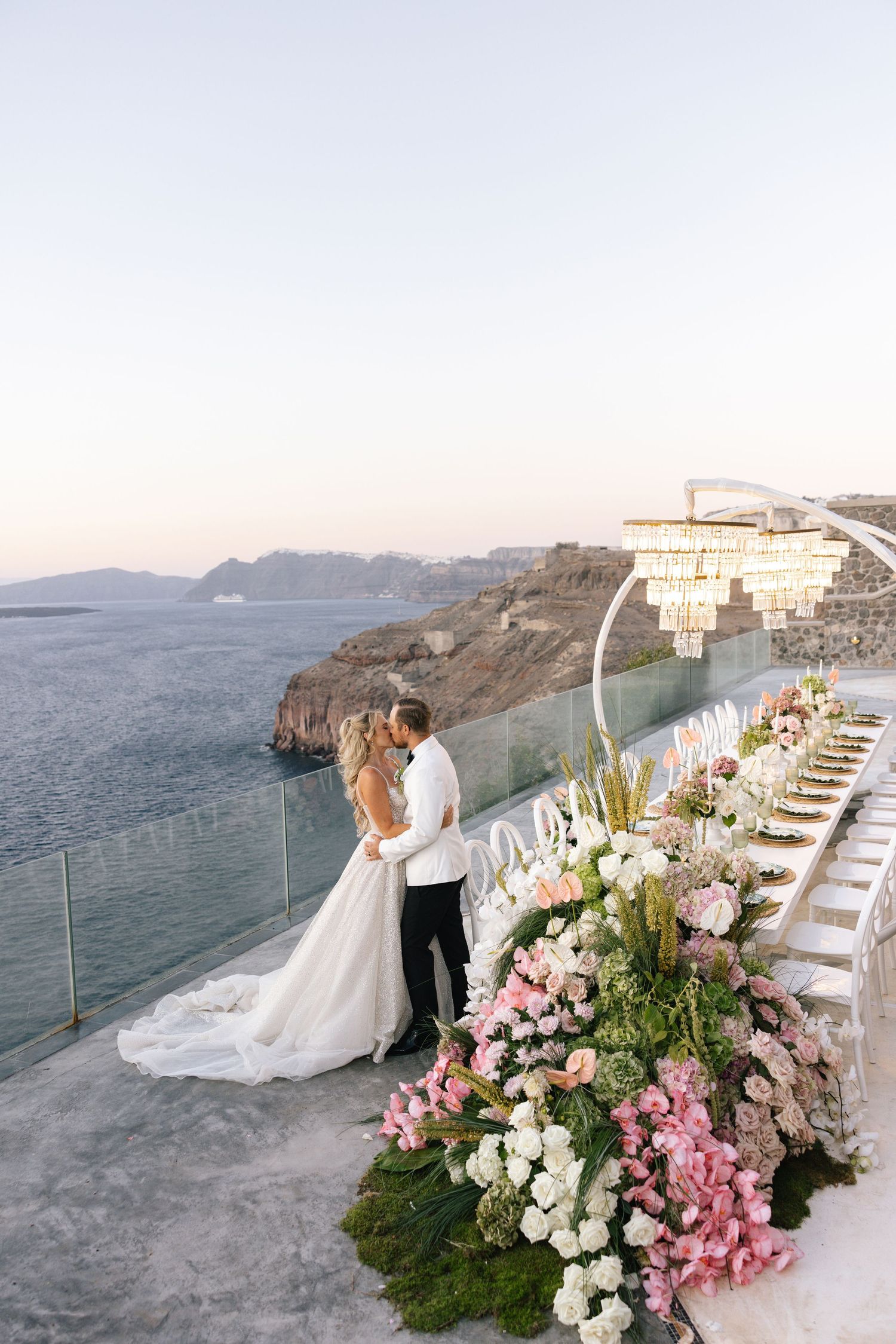 Bride and groom embrace on a cliffside terrace in Santorini with pink floral arrangements and ocean views.
