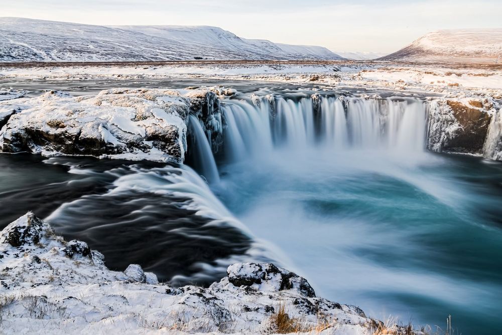 Godafoss waterfall frozen in winter with snow-covered rocks and turquoise rushing water in Iceland.