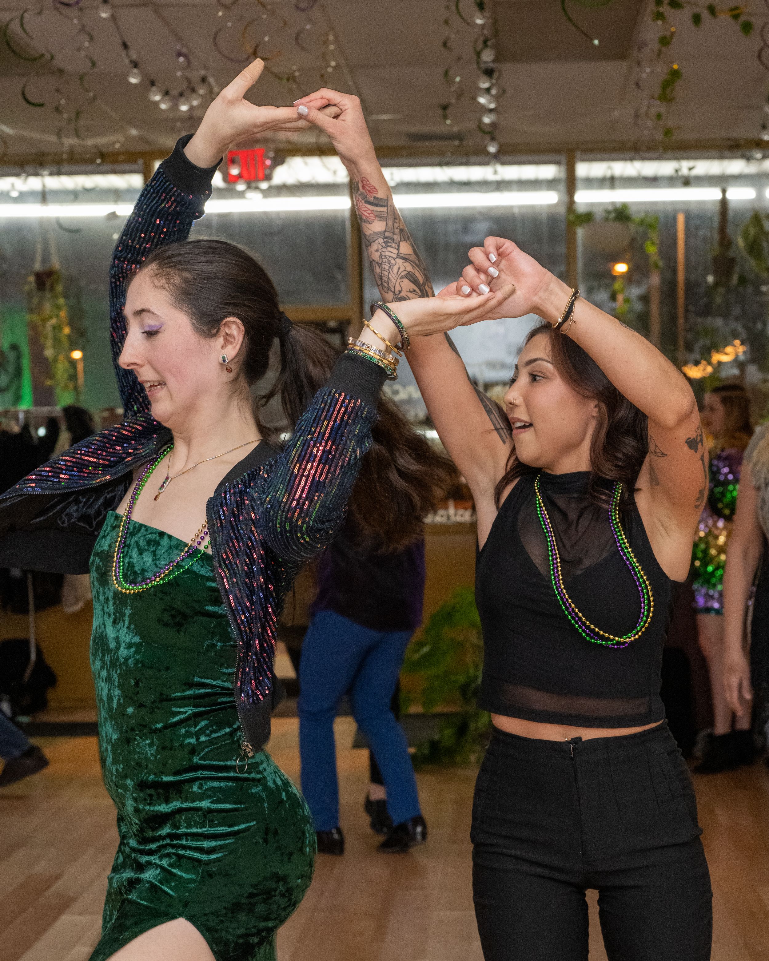 Two women dancing energetically at a Mardi Gras party wearing green velvet dress and black crop top with beaded necklaces.