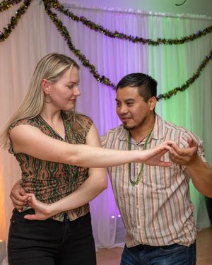 A man and woman dancing together at a festive event with green garland decorations and colorful lights.