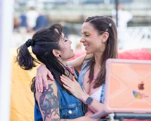 Two women embrace and smile at each other at a Pride event, one with tattoos and rainbow eye makeup.