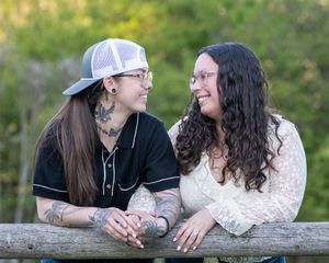 Two women with tattoos smiling at each other while leaning on a wooden fence outdoors.