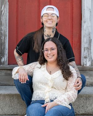 Two smiling women pose on steps in front of a red barn door, one wearing a black shirt and cap, the other in a lace blouse.