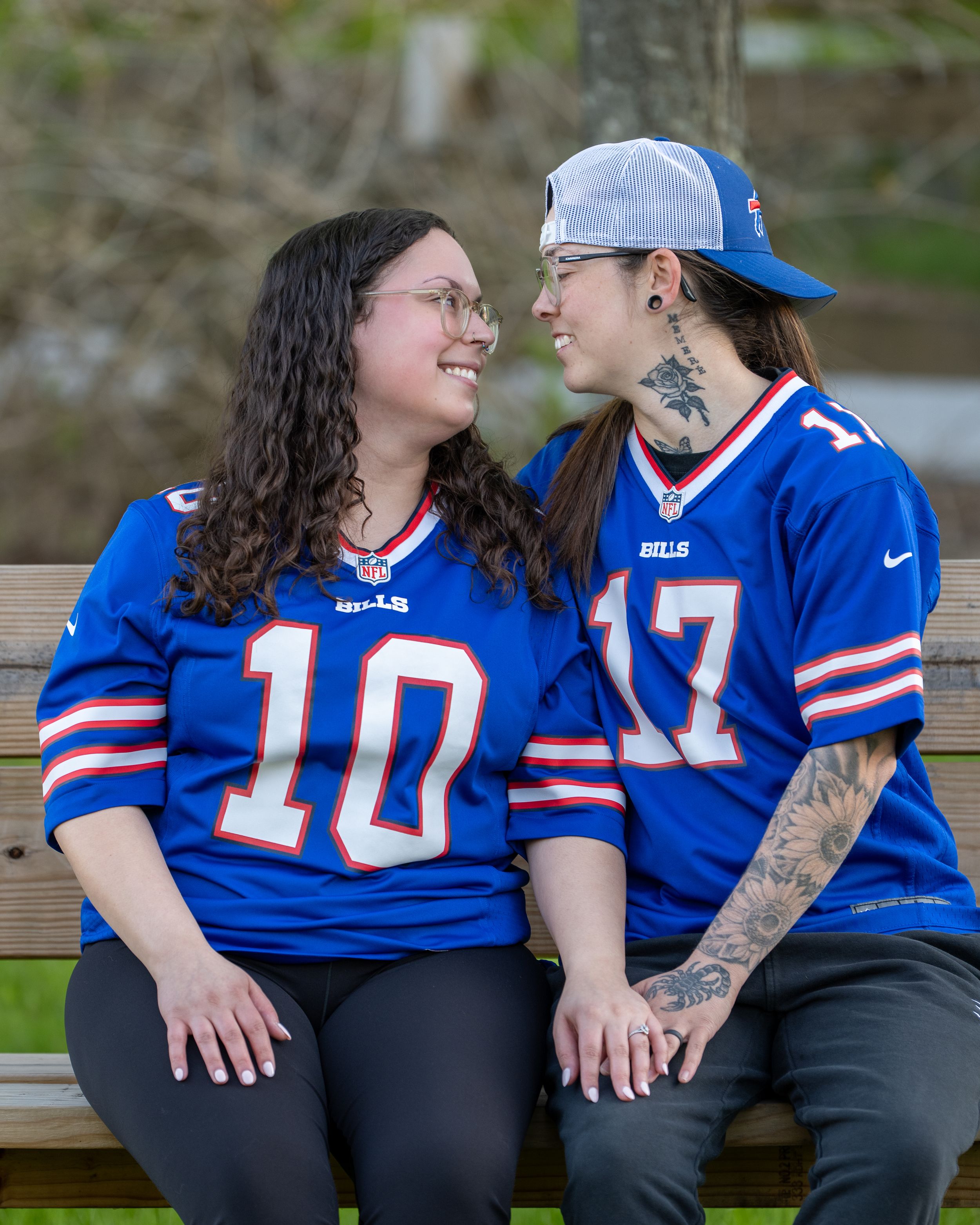 Two women in Buffalo Bills jerseys #10 and #17 sit on a bench smiling at each other.