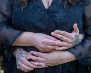 Close-up of two people embracing, showing tattooed hands and rings, wearing a black floral embroidered dress.