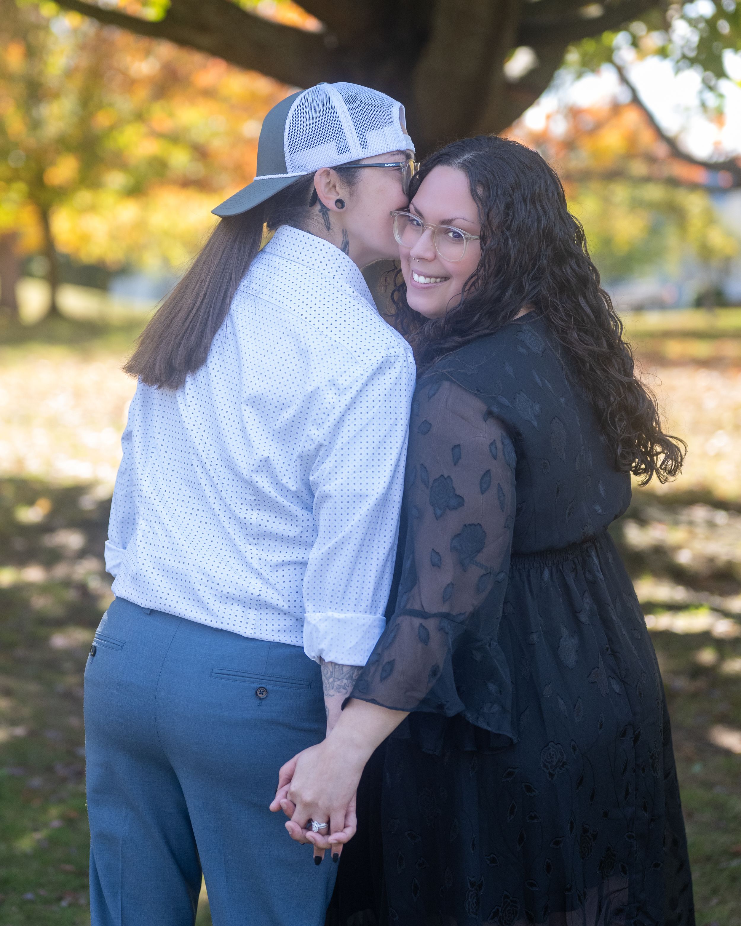 Two women holding hands and sharing a kiss outdoors in a park with colorful autumn foliage.