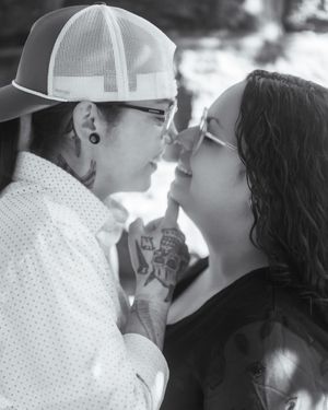 Black and white photo of two people with tattoos and glasses sharing a tender moment, nearly kissing outdoors.