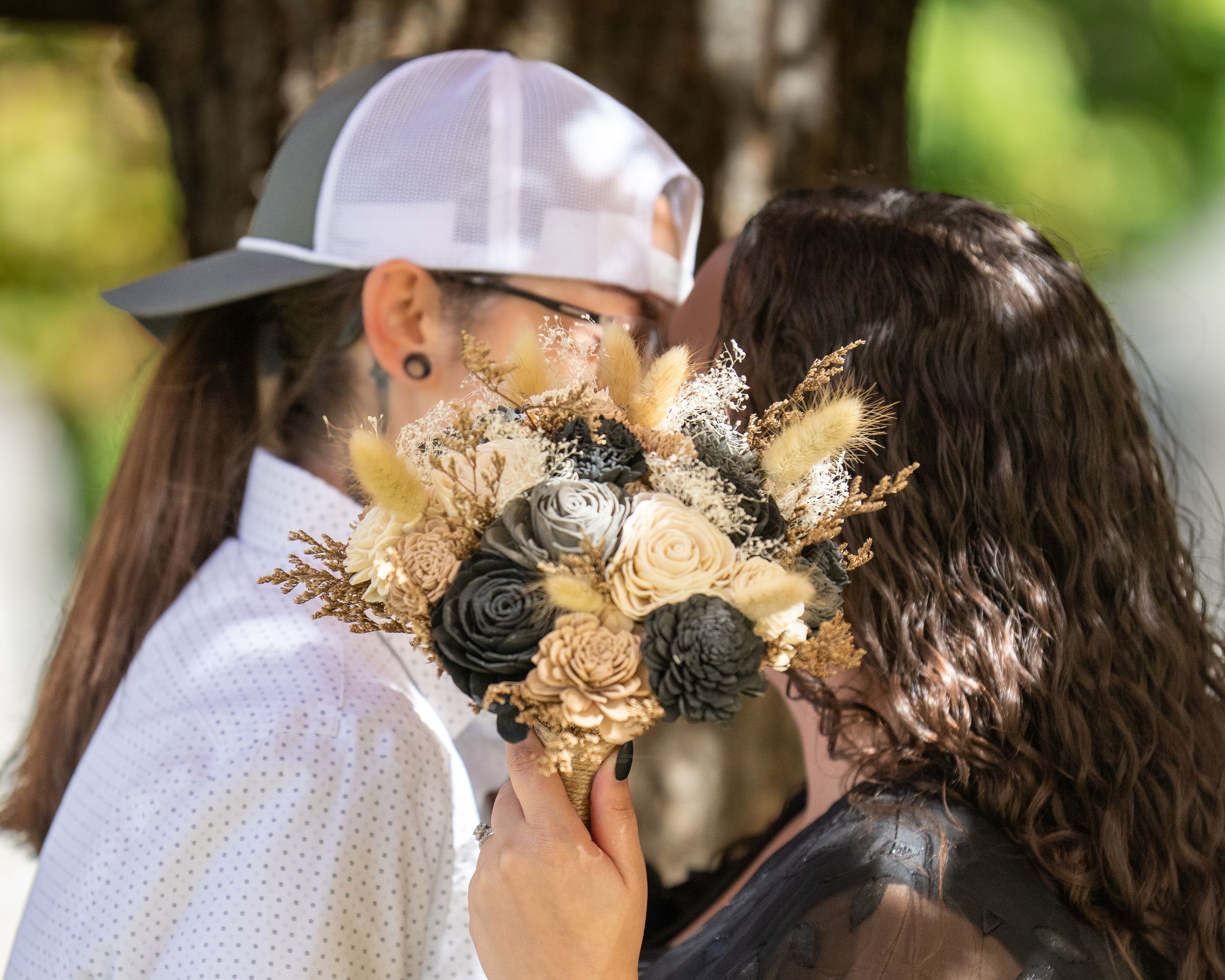 Couple sharing a kiss behind a rustic bouquet of dried flowers at an outdoor wedding ceremony.