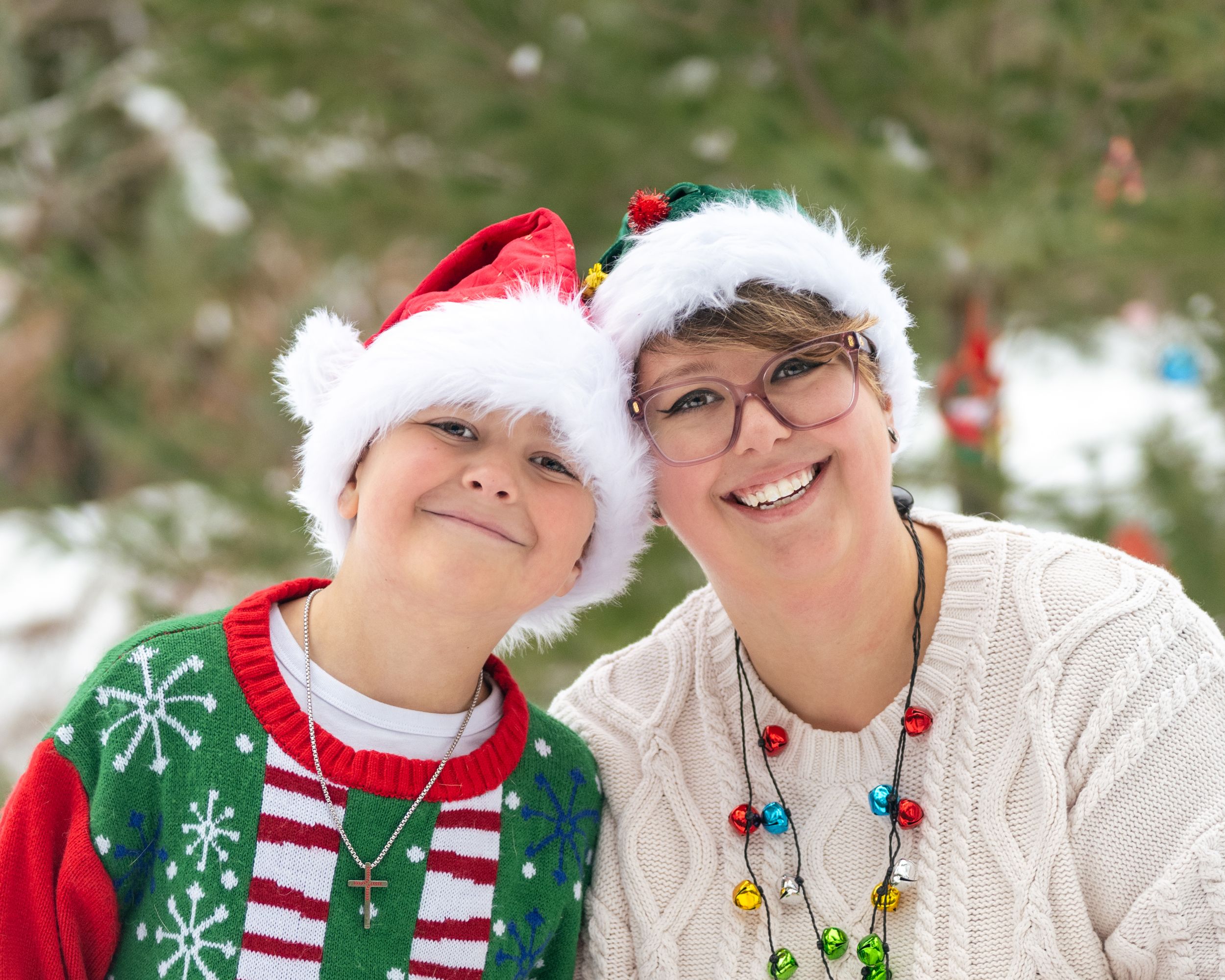 Two people wearing Santa hats and festive sweaters smile together outdoors in a snowy winter holiday setting.