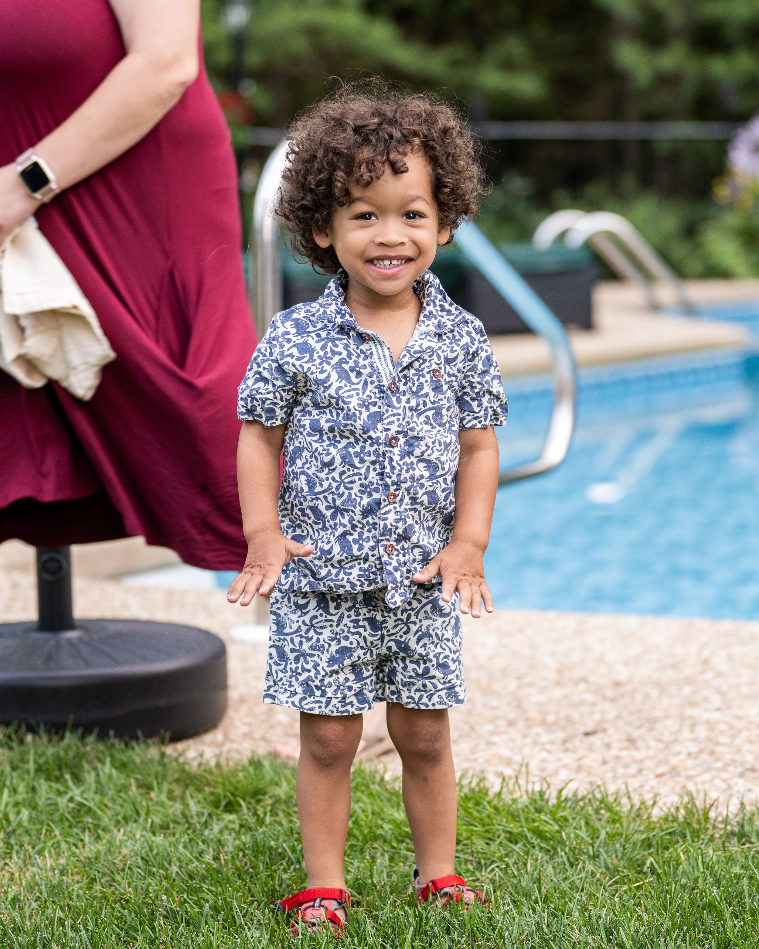 Toddler boy smiling in navy floral shirt and shorts with red sandals, standing on grass near a pool.