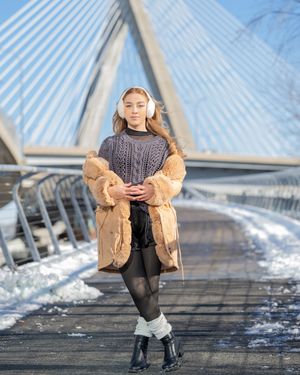 Young woman in fur coat, cable-knit sweater, and ear muffs poses on snowy bridge walkway in winter.