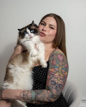 Woman with colorful tattoo sleeve holding a fluffy Ragdoll cat while playfully sticking out her tongue.
