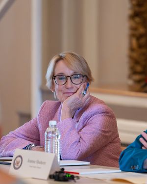 Woman in pink blazer sits at conference table with nameplate, water bottle, resting chin on hand thoughtfully.