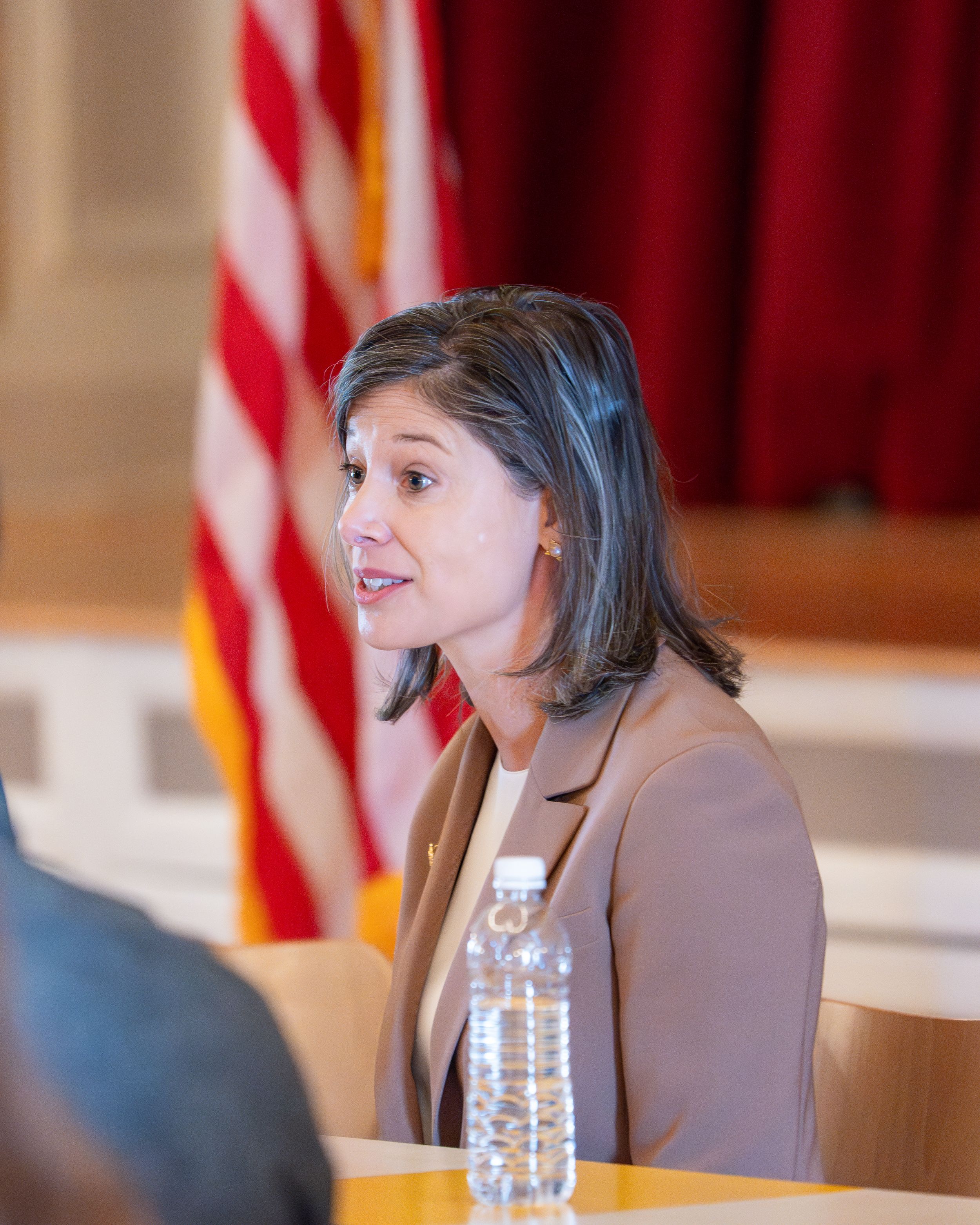 Woman in tan blazer speaking at a table with an American flag in the background.