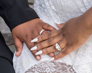 Close-up of newlywed couple holding hands showing wedding rings, bride wearing white lace dress with long white nails.