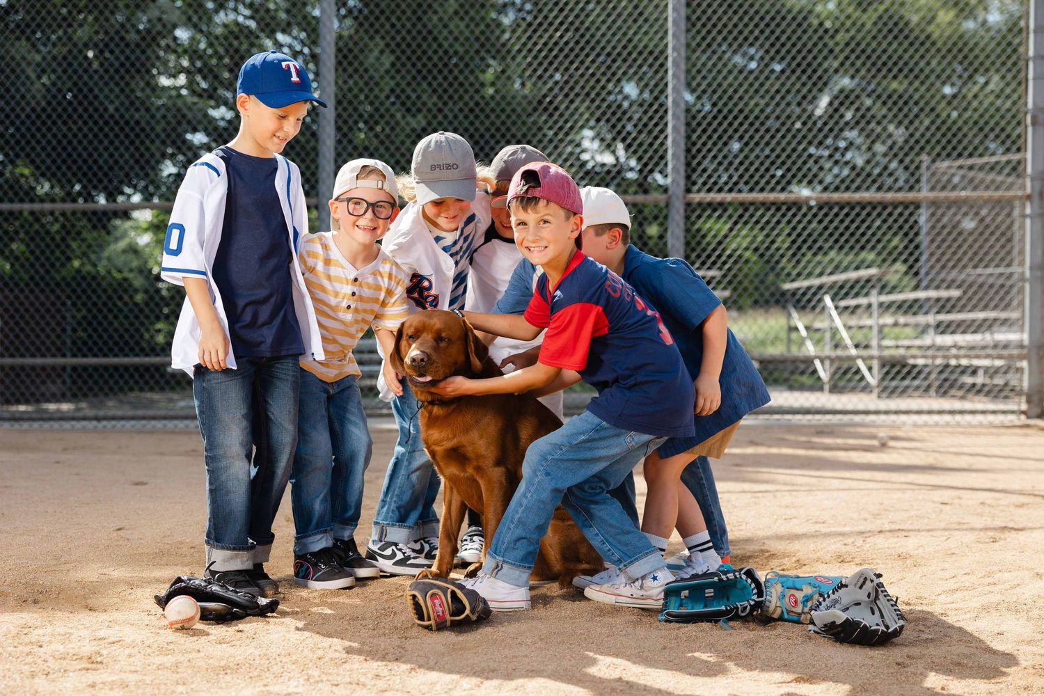 Group of young boys in baseball uniforms petting a brown dog on a dirt baseball field.