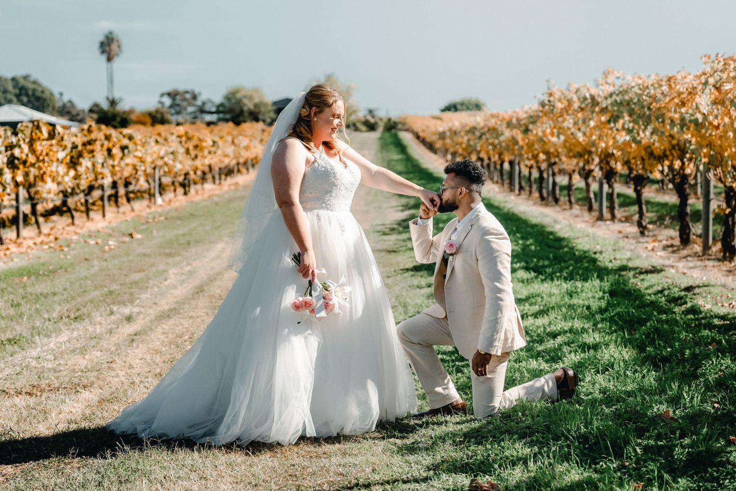 Bride in white gown and groom in beige suit share romantic moment in autumn vineyard rows on sunny wedding day.