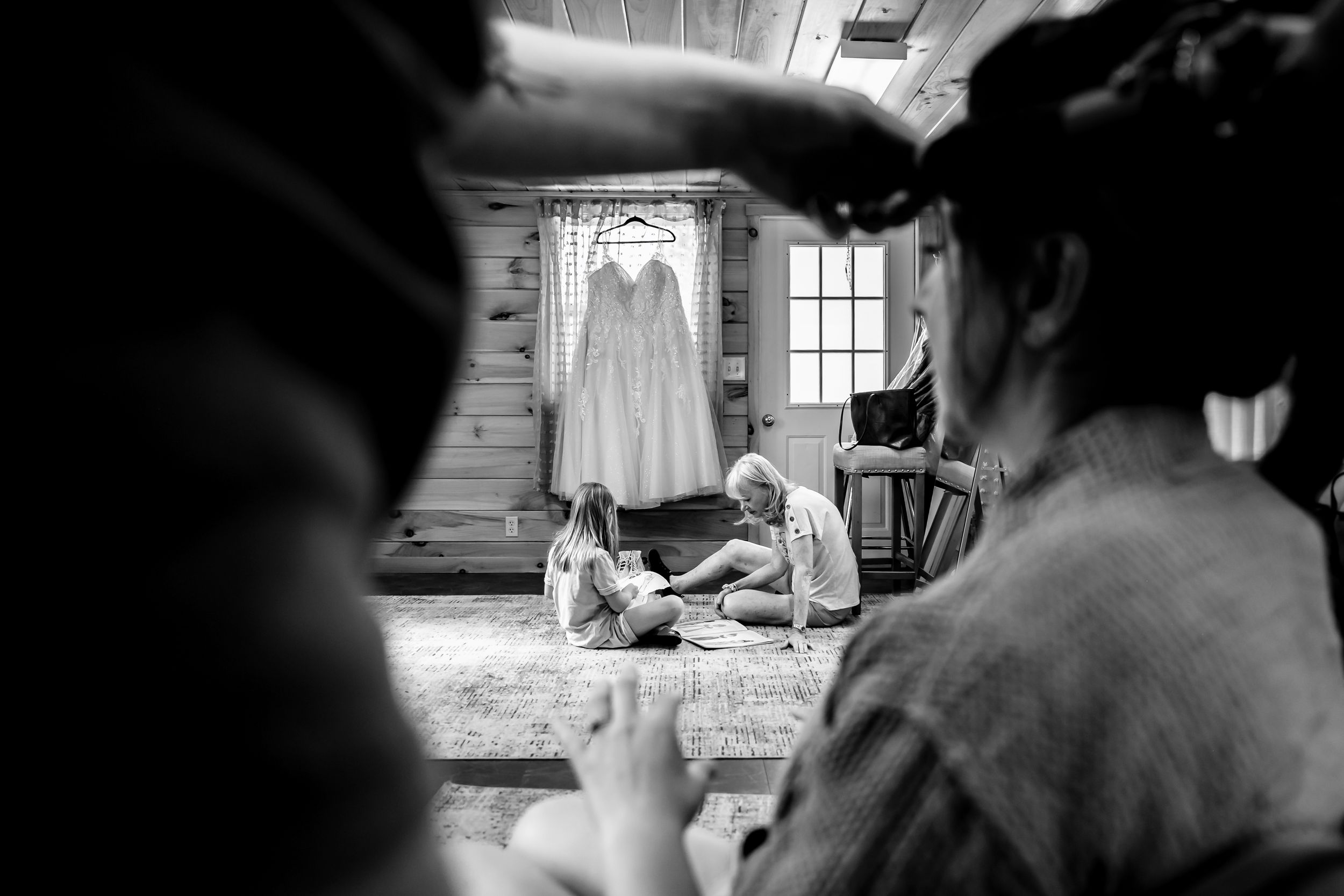 Black and white photo of two people sitting on floor before a wedding dress hanging in a rustic cabin window.
