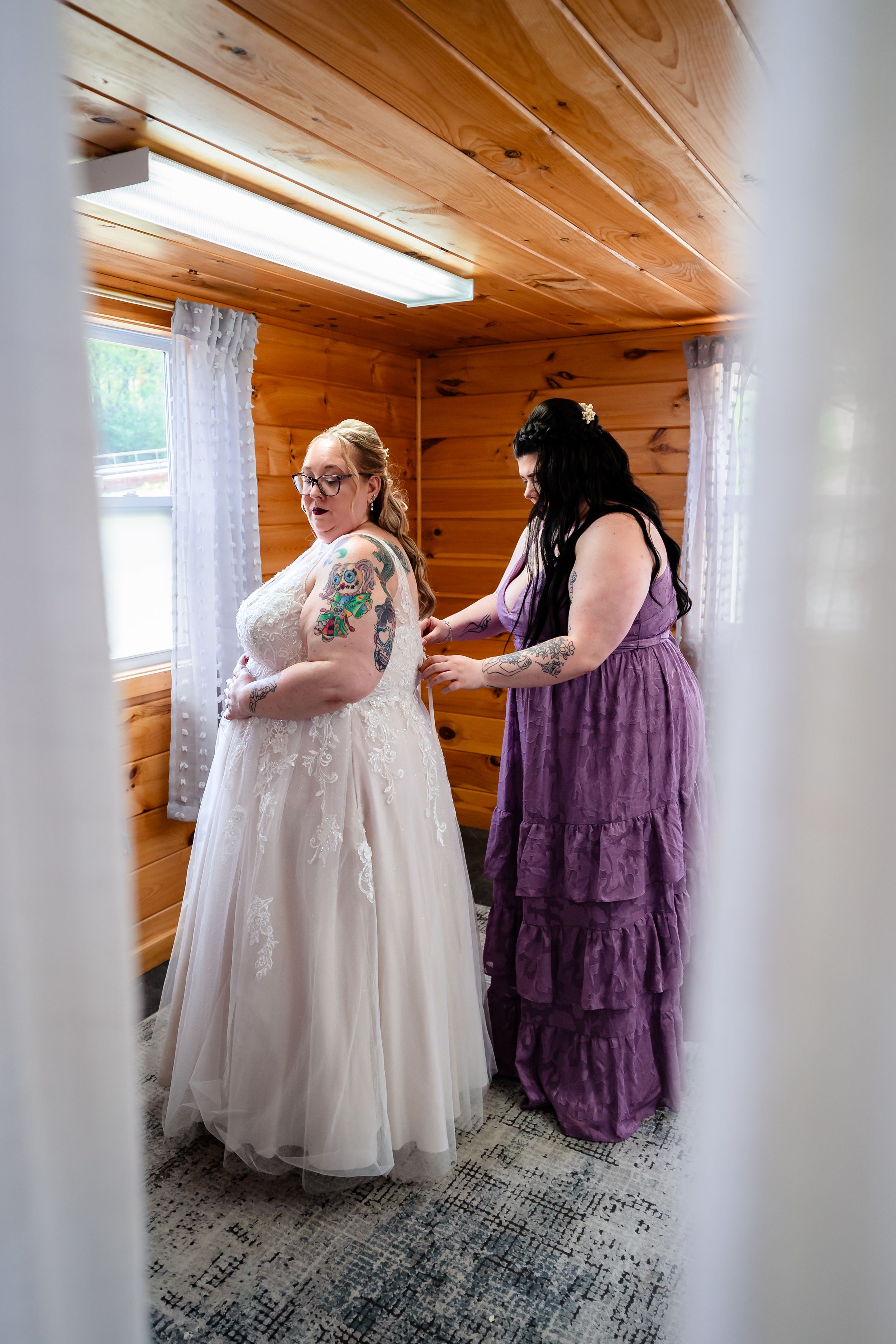 Bride in white lace gown being helped by bridesmaid in purple dress inside rustic wooden cabin.