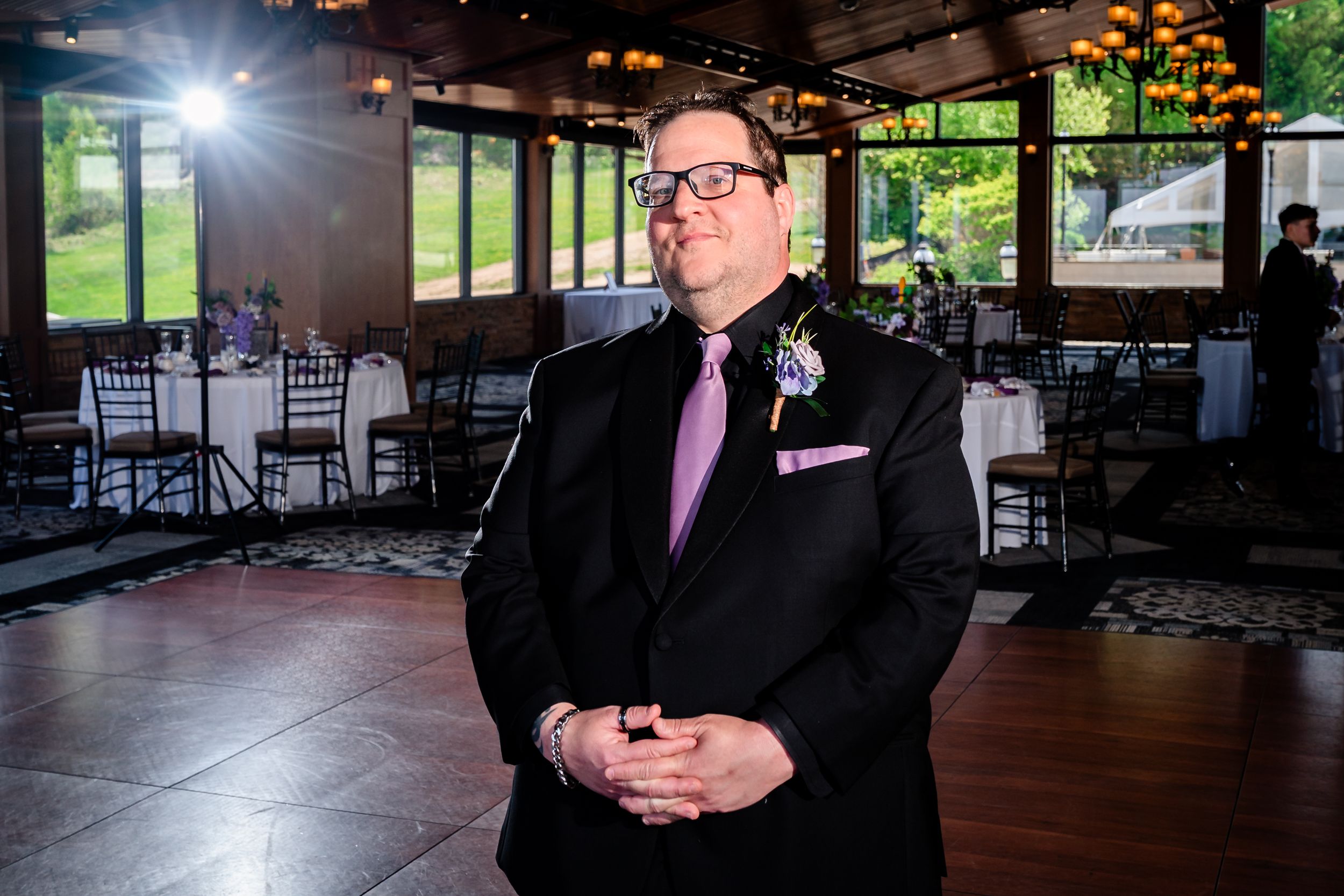 Groom in black suit with purple tie and boutonniere stands in elegant wedding reception venue.
