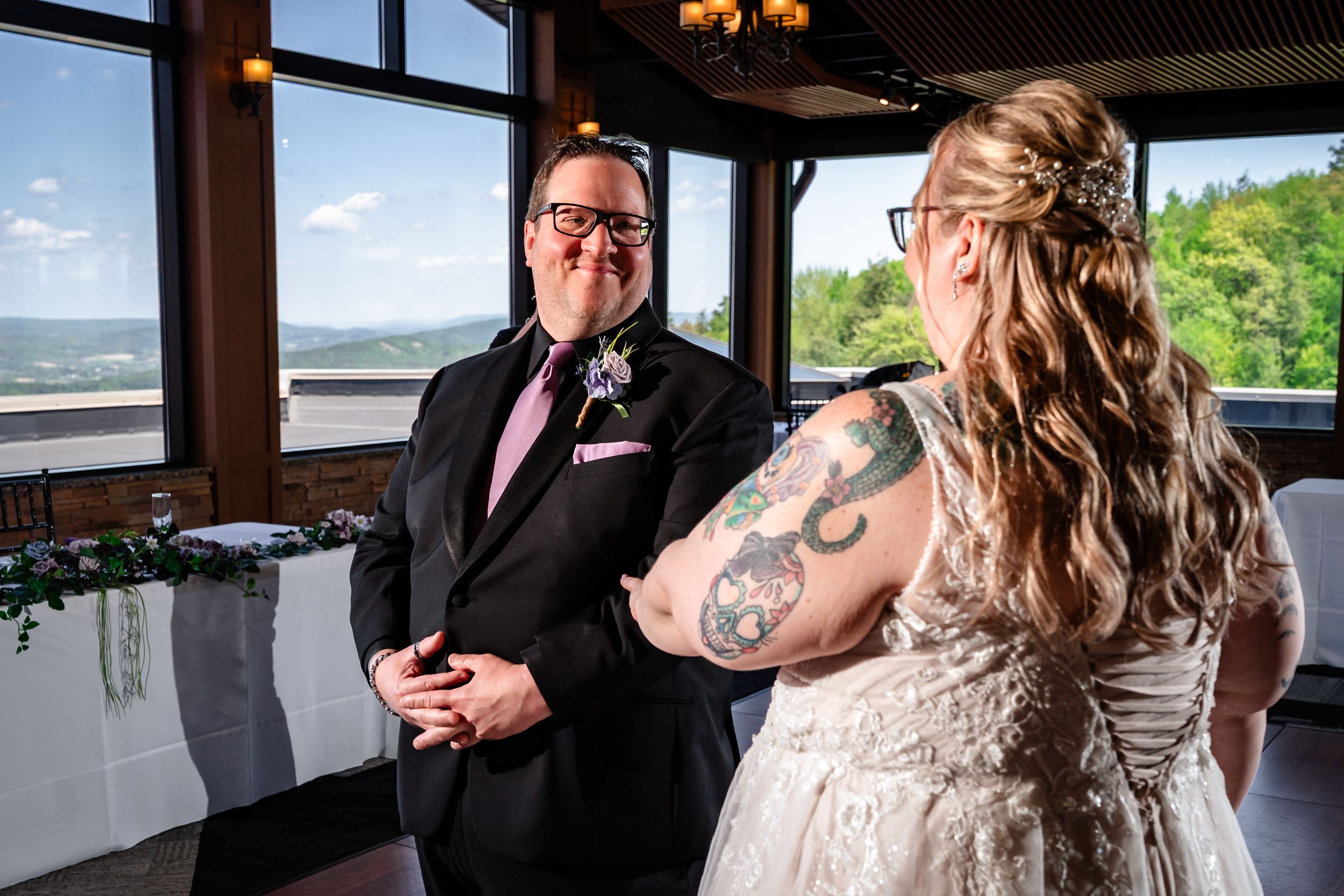 Groom in black suit smiling at tattooed bride in white wedding gown during first look moment.