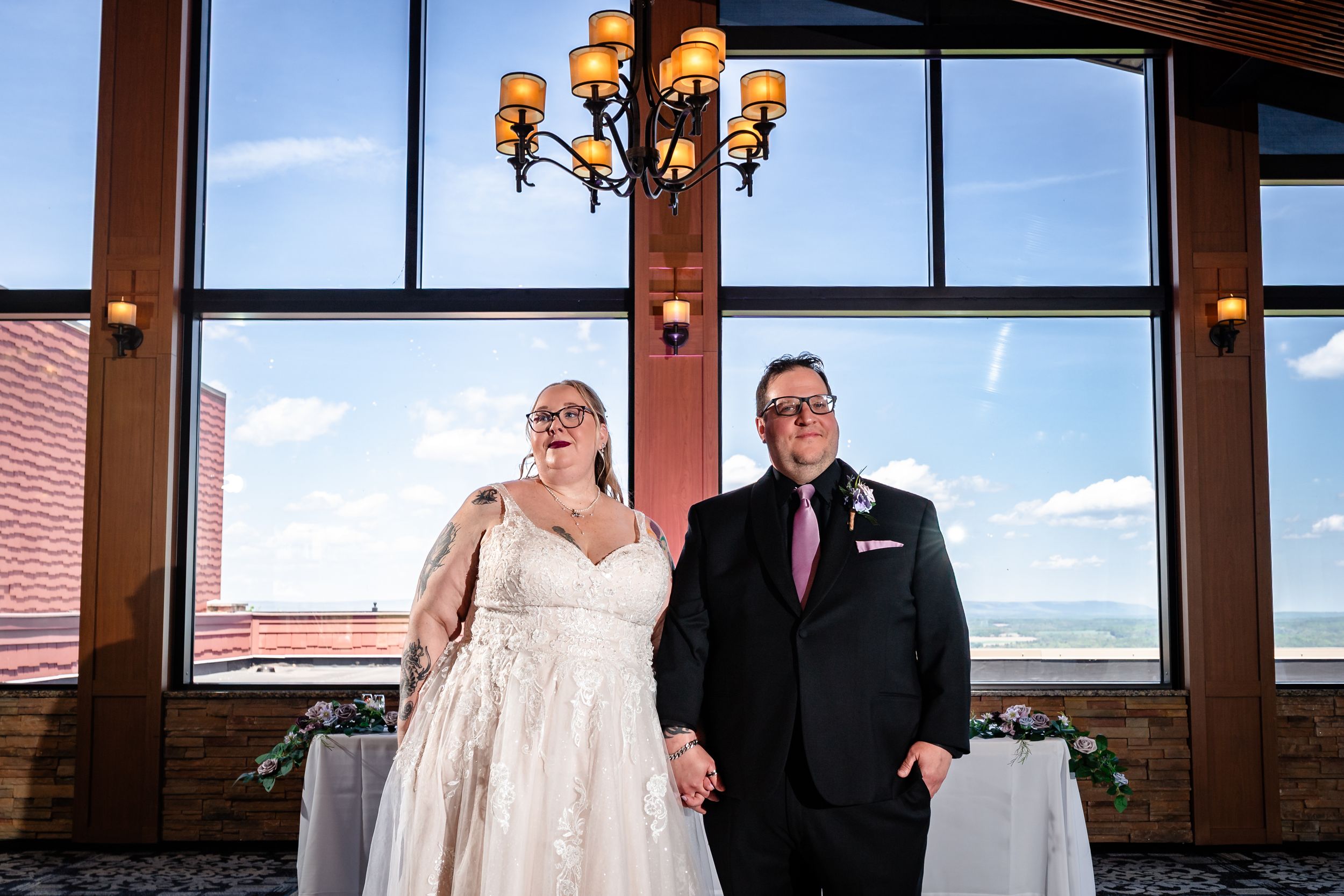 Bride and groom hold hands smiling under chandelier at elegant wedding venue with large windows.