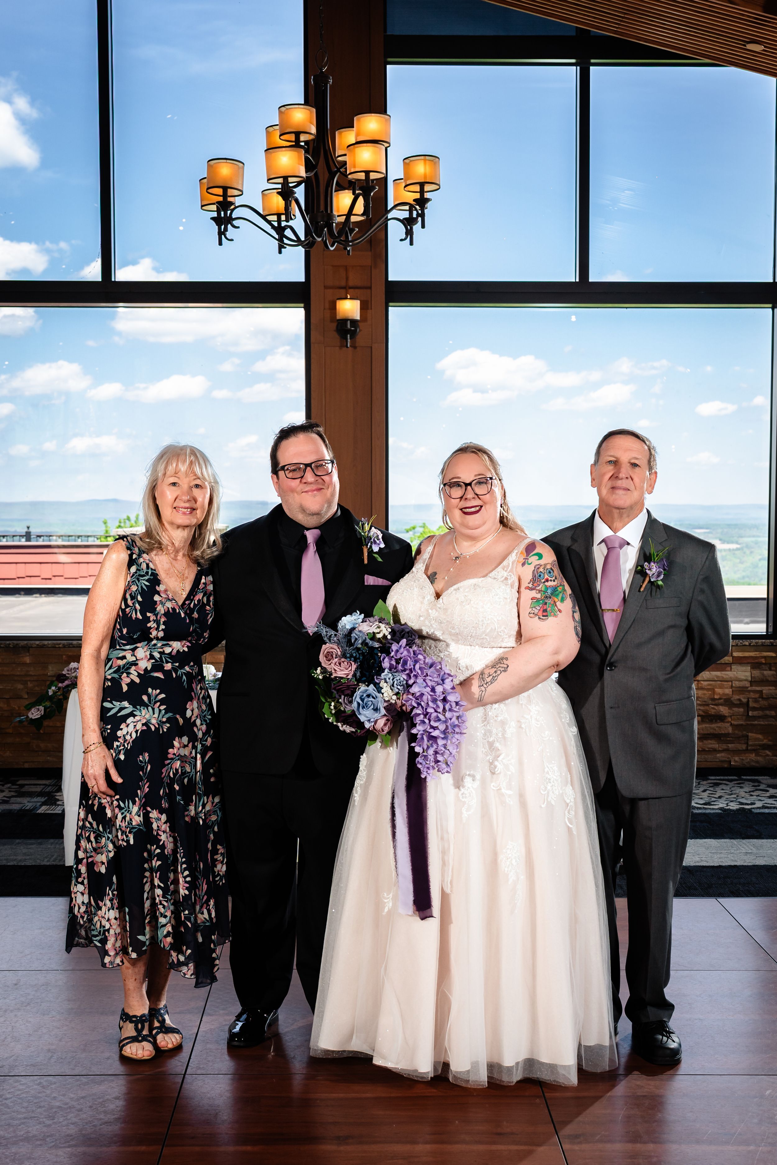 Wedding party of four posing indoors with chandelier and large windows showing blue sky behind them.