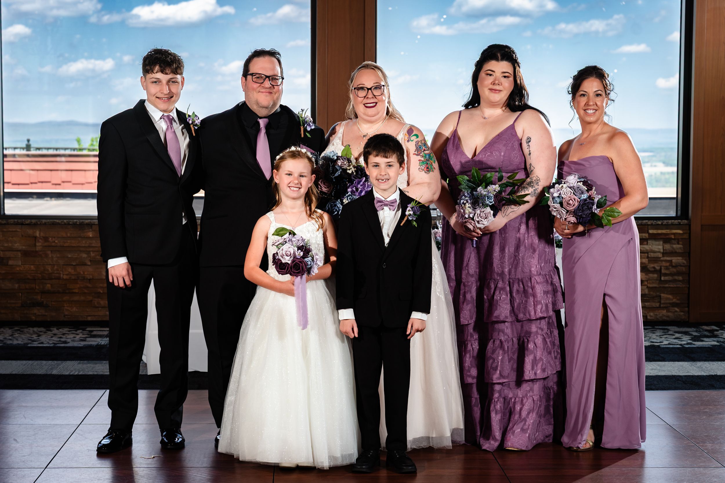 Wedding party poses together in purple dresses and suits at indoor venue with scenic windows.