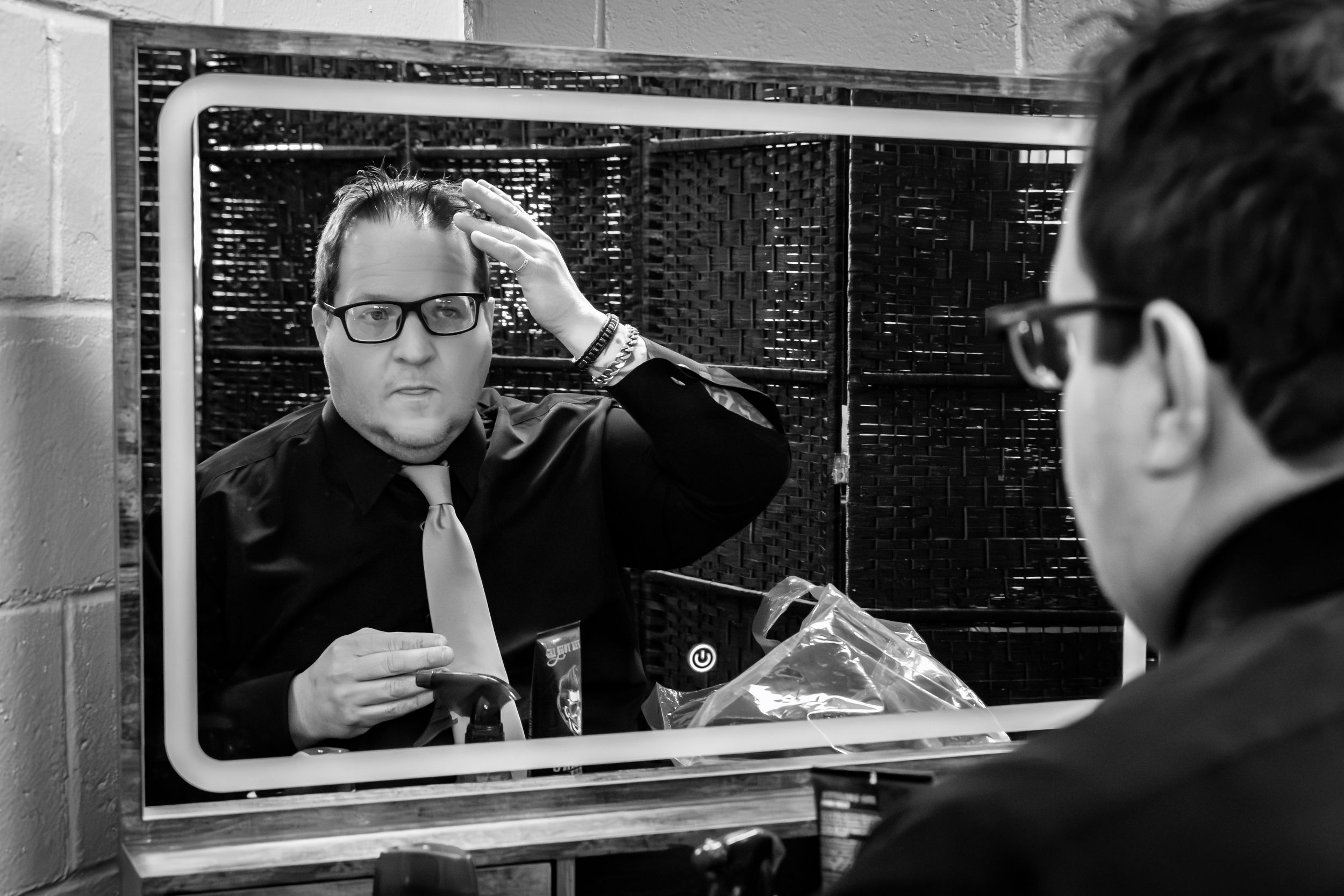 Black and white photo of a man fixing his hair while looking in a mirror, wearing glasses and a dark suit.