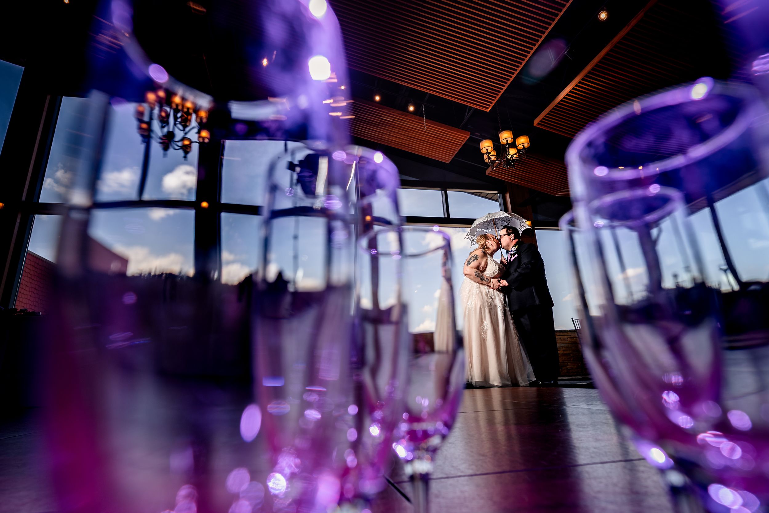 Bride and groom sharing romantic kiss framed by purple glowing champagne flutes at wedding reception.