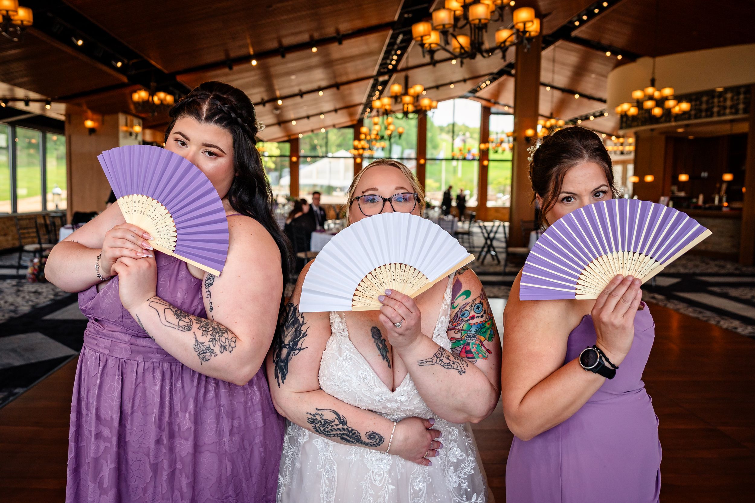 Bride and three bridesmaids in purple dresses playfully hiding faces behind decorative fans at wedding.