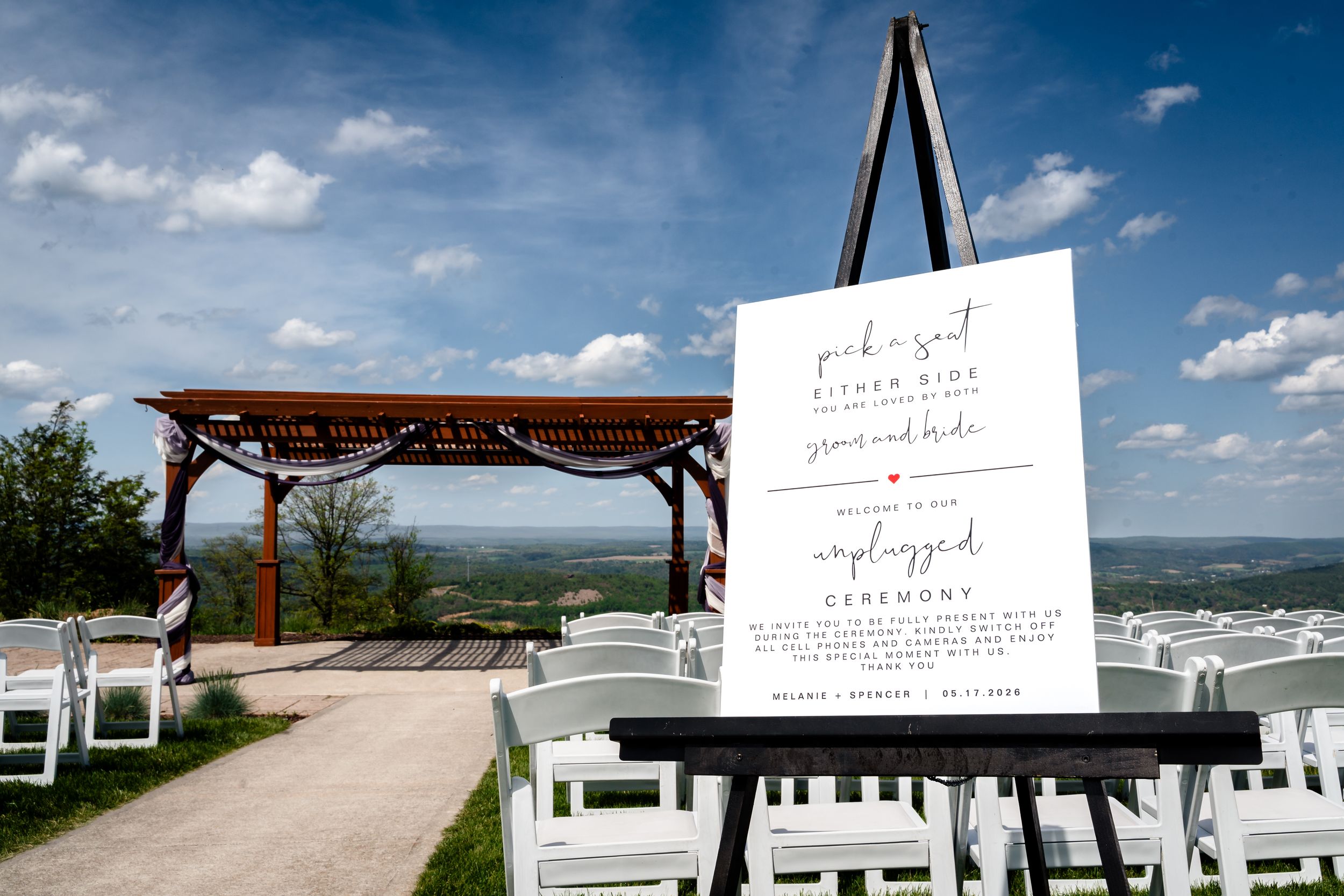 Outdoor wedding ceremony sign announcing unplugged ceremony with white chairs and scenic valley backdrop.