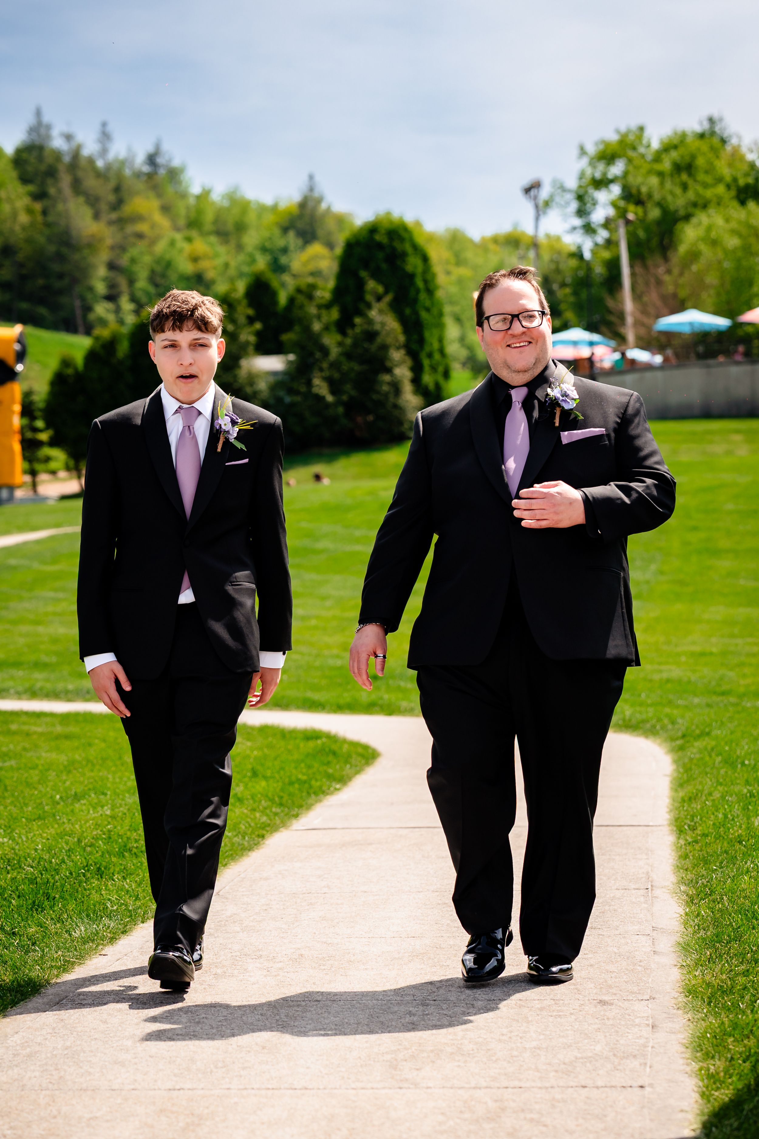 Two groomsmen in black suits with lavender ties walk along a winding path at an outdoor wedding venue.