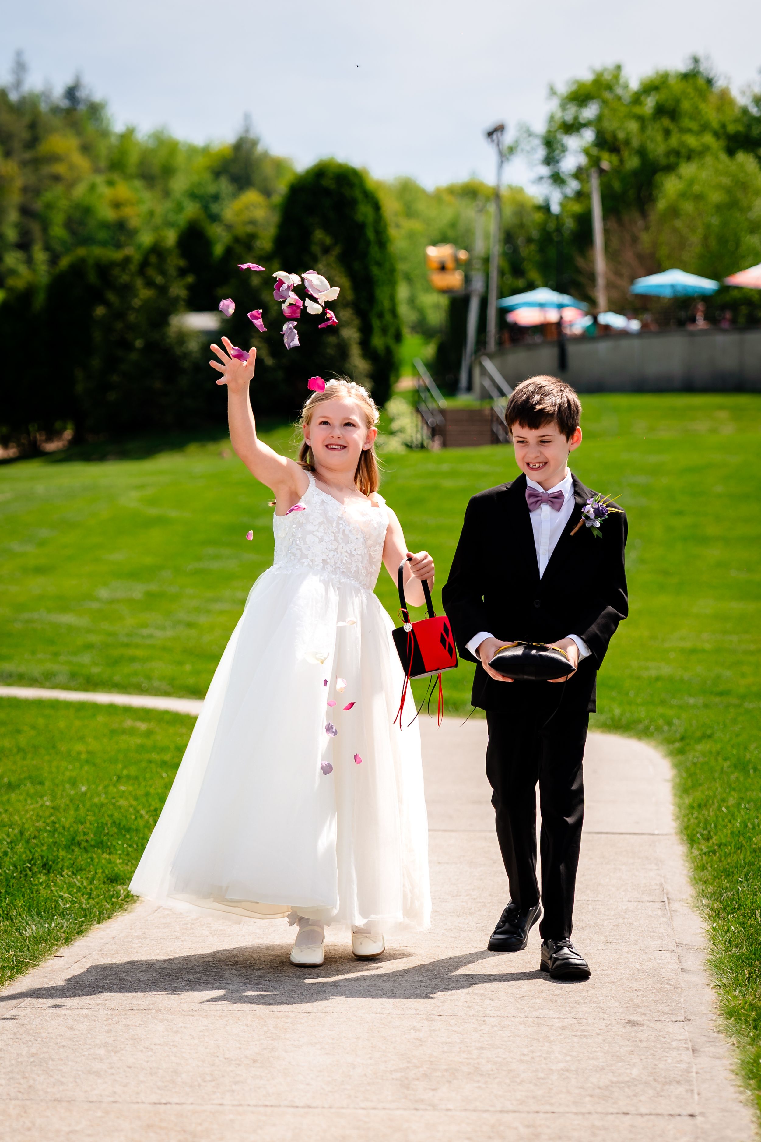 A flower girl in white dress tosses petals while a ring bearer in black suit walks beside her on a garden path.