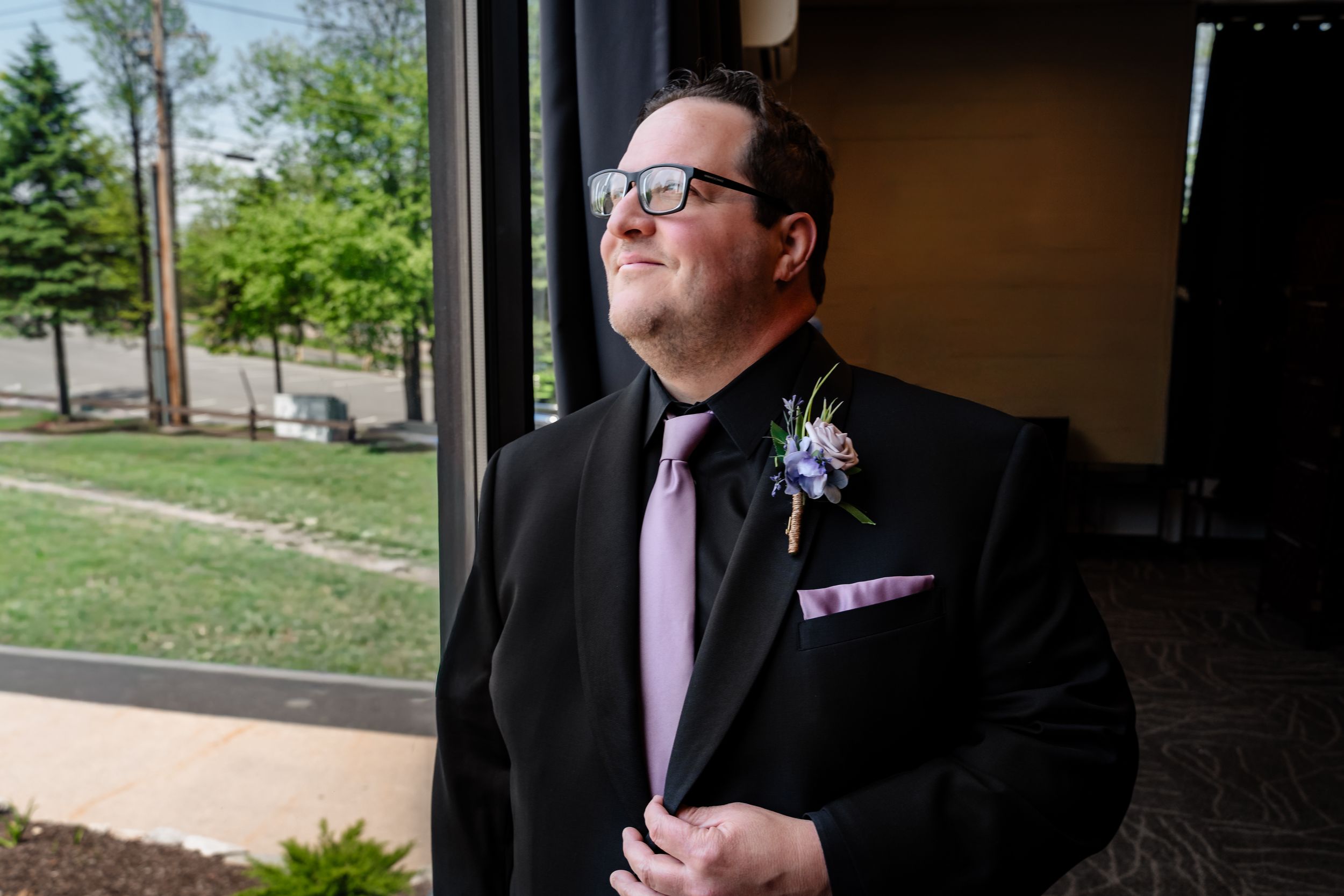 Groom in black suit with pink tie and boutonniere gazing upward near large window on wedding day.