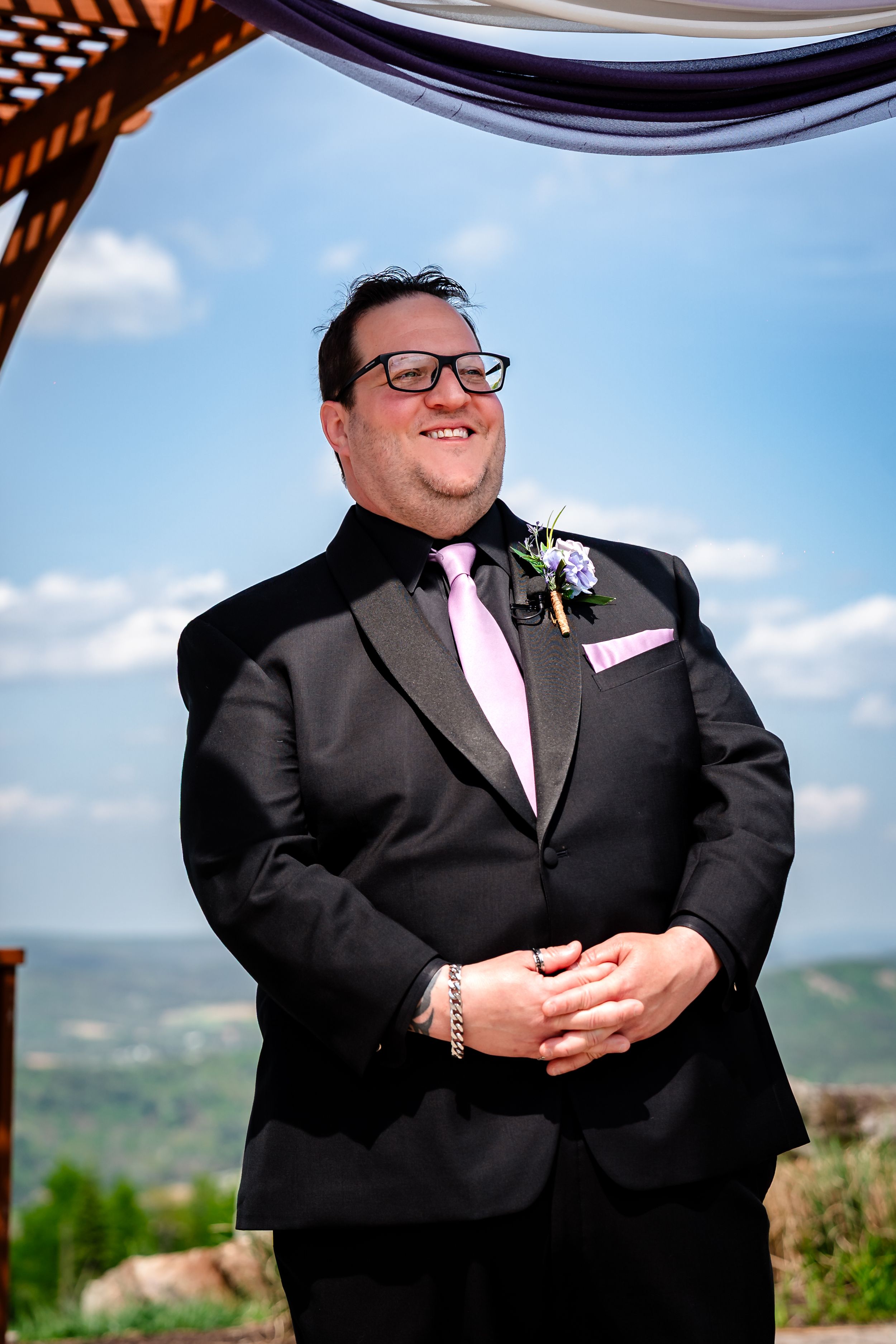 Smiling groom in black tuxedo with pink tie and boutonniere stands at outdoor wedding ceremony under blue sky.