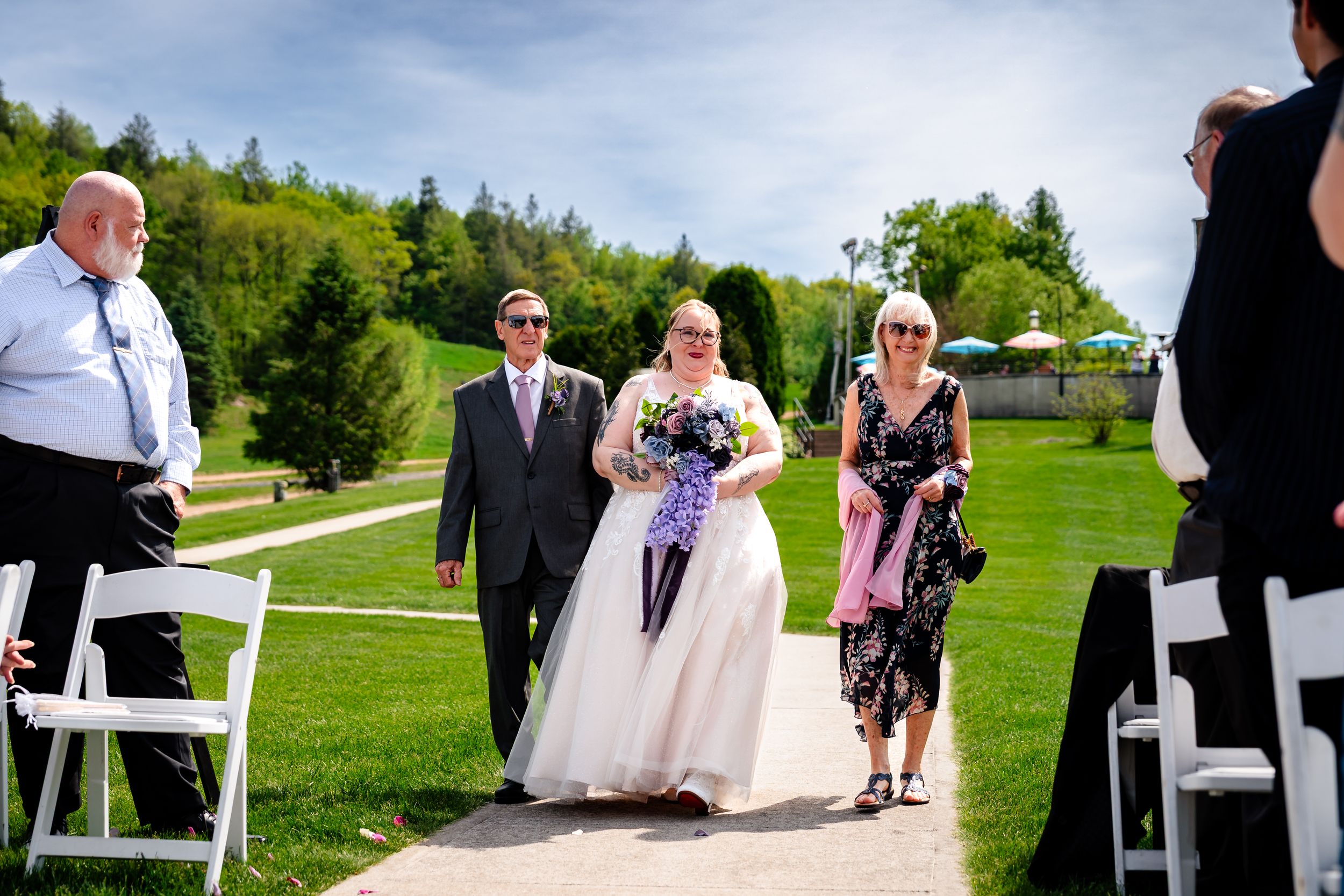Bride walking down outdoor aisle with escorts at sunny wedding ceremony, white chairs and green hills in background.