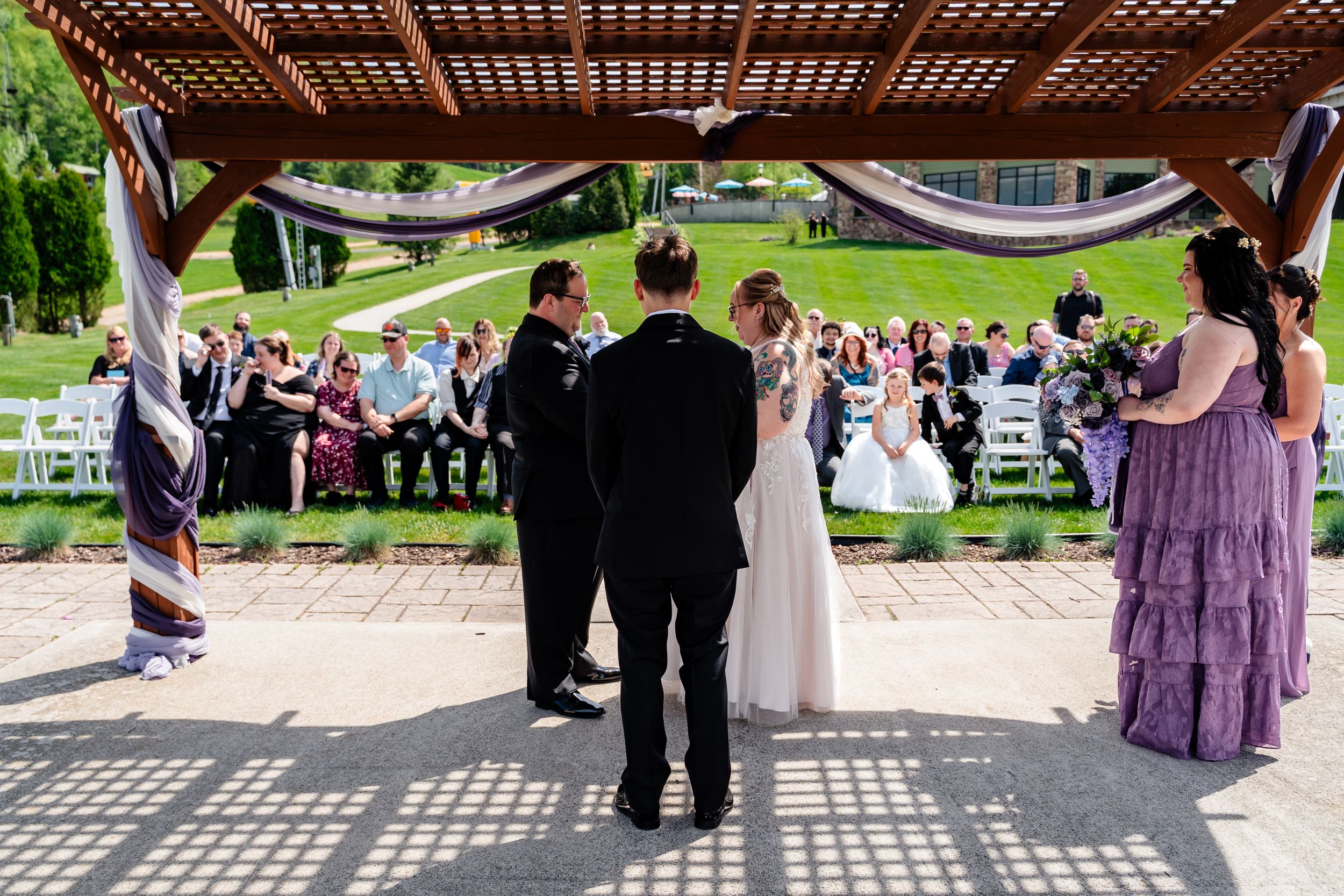 Wedding ceremony under wooden pergola with bride, groom, and officiant surrounded by guests outdoors.