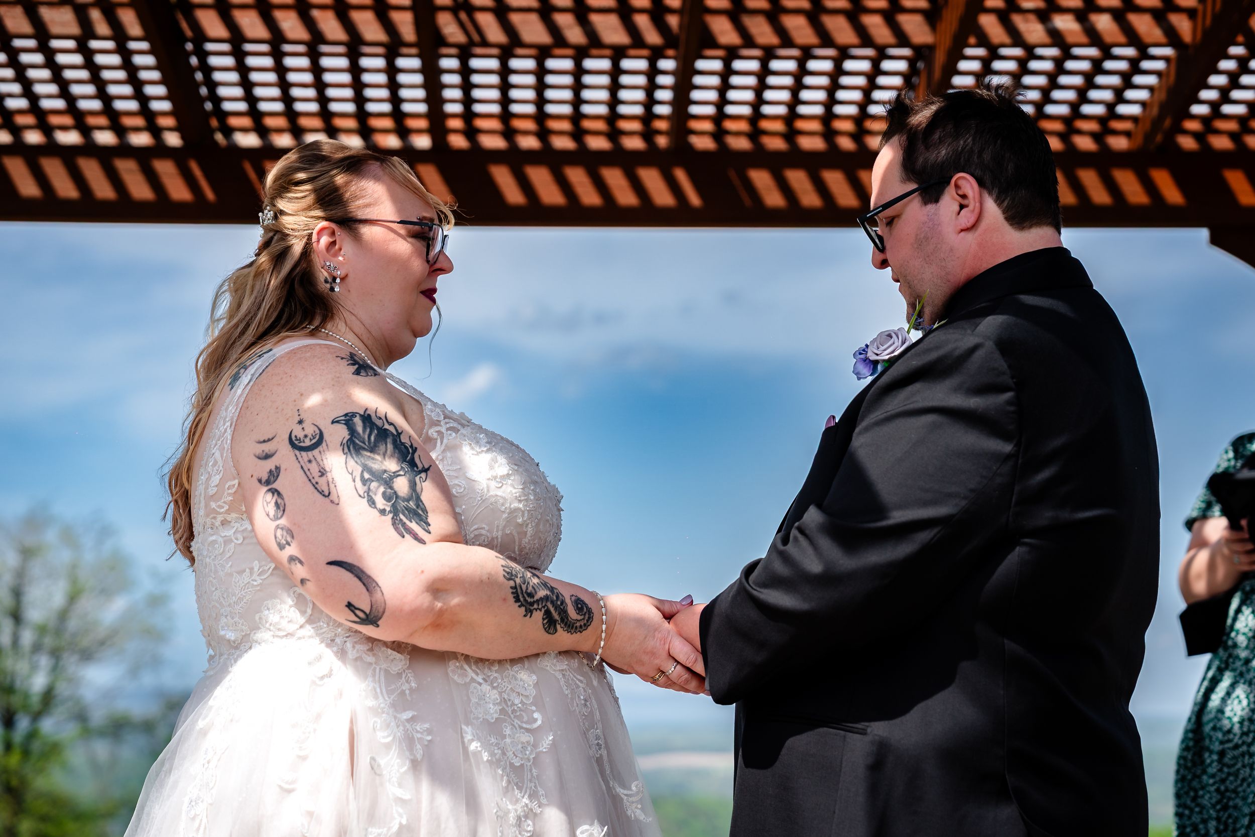 Couple exchanging vows under a wooden pergola, bride with tattoos in white gown, groom in black suit.