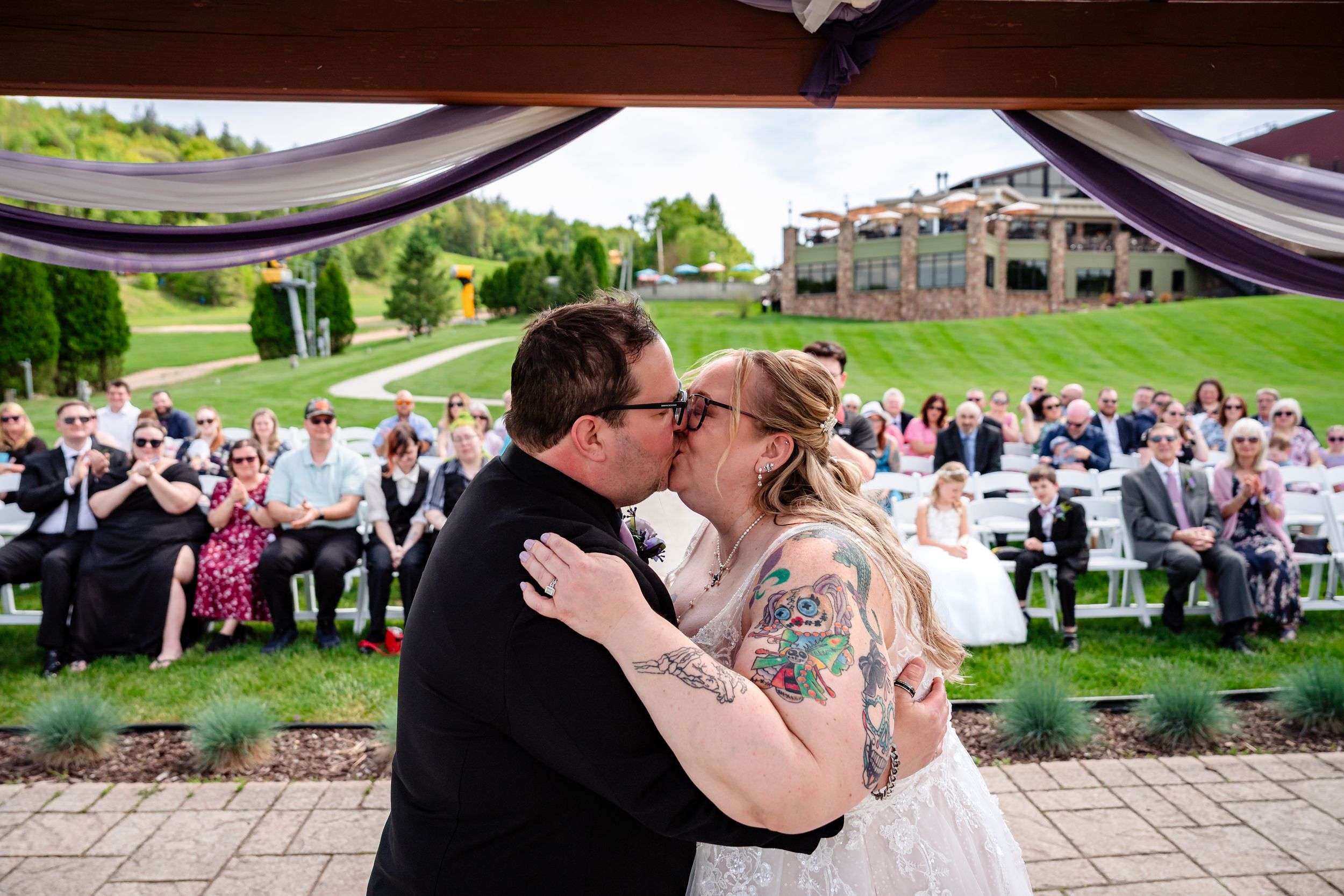 Bride and groom sharing first kiss under pergola with guests applauding behind them.