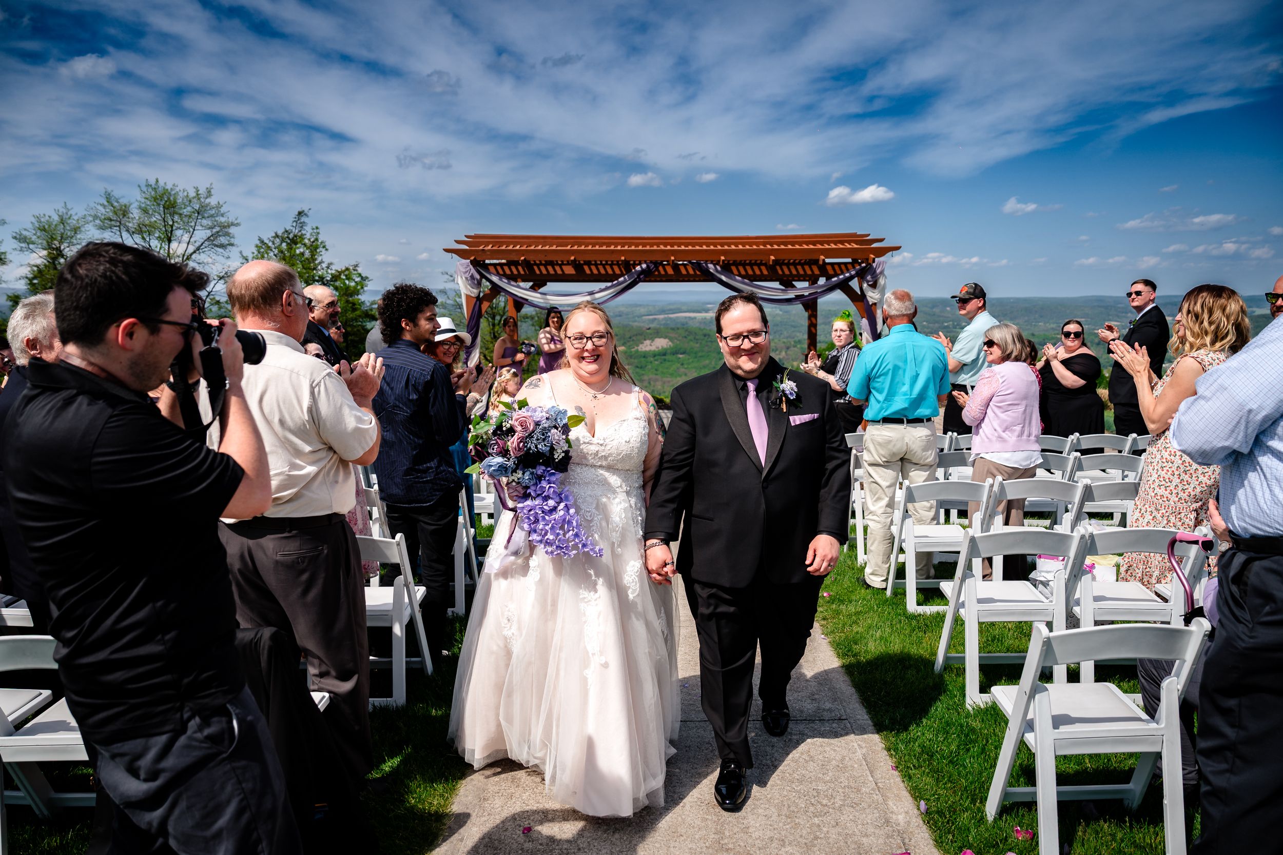 Bride and groom walk back down aisle after outdoor wedding ceremony under blue sky with guests applauding.