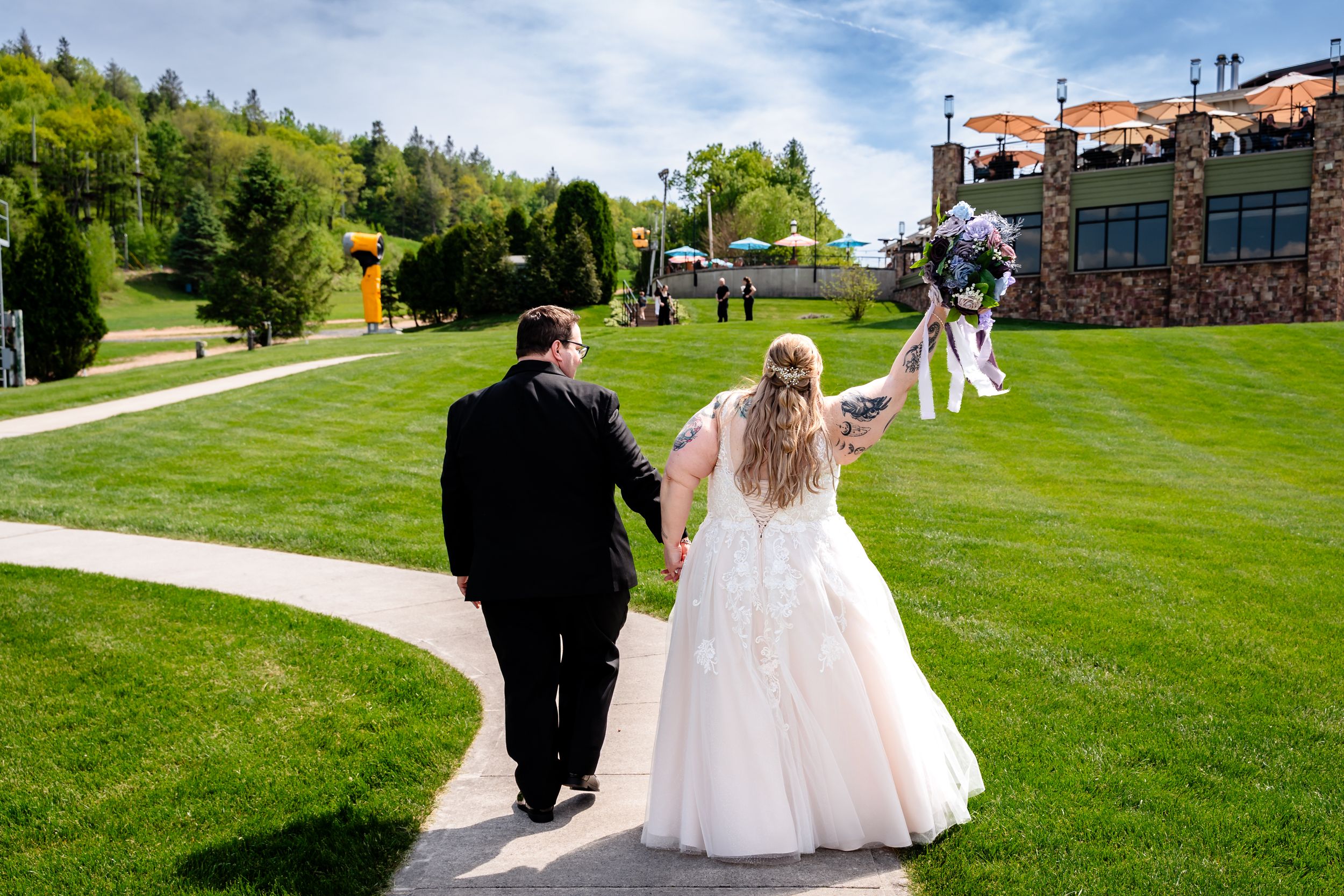 Bride and groom walk hand-in-hand along a garden path at their wedding venue on a sunny day.