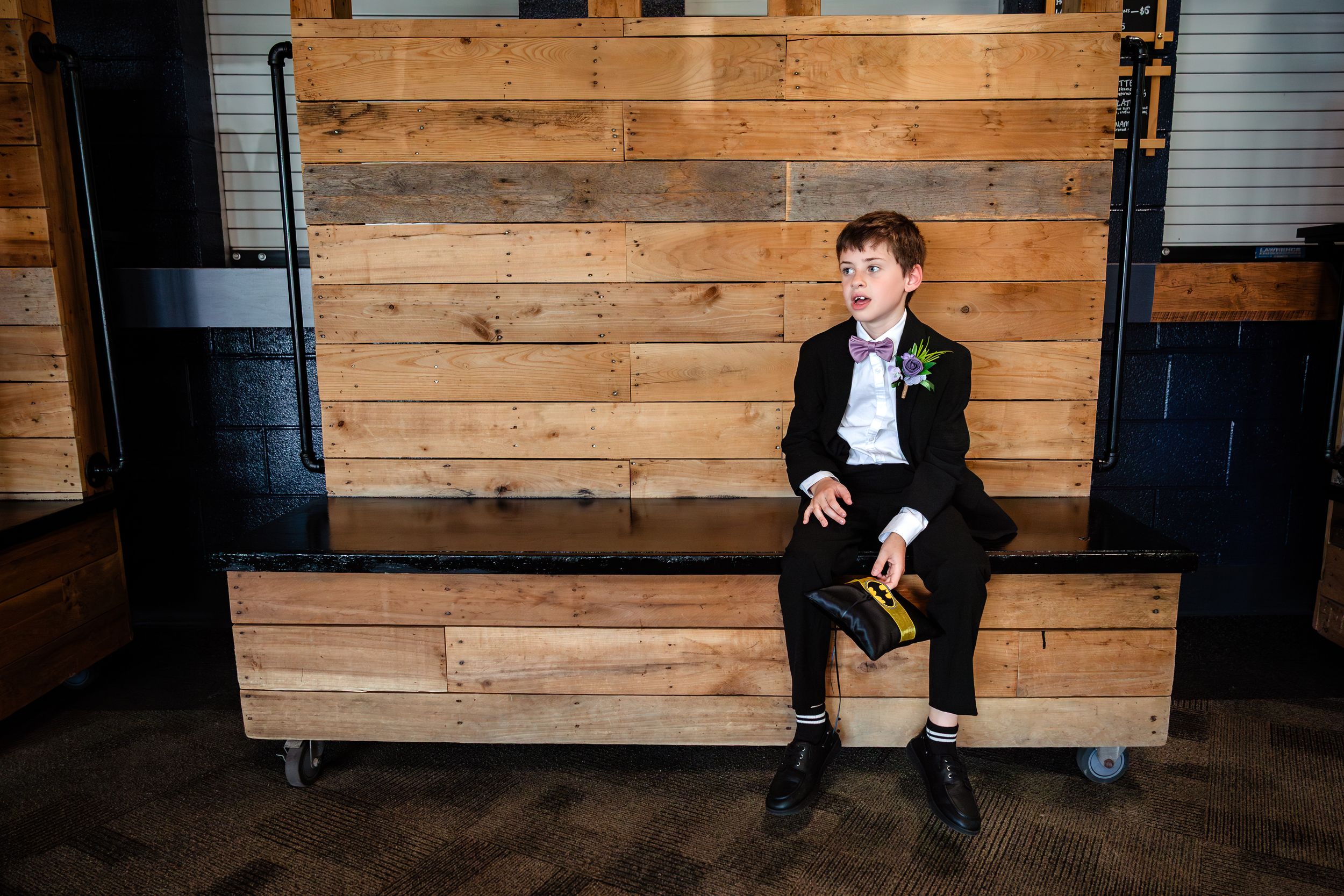 Young ring bearer in black suit sits alone on rustic wooden bench holding a small hat at wedding reception.