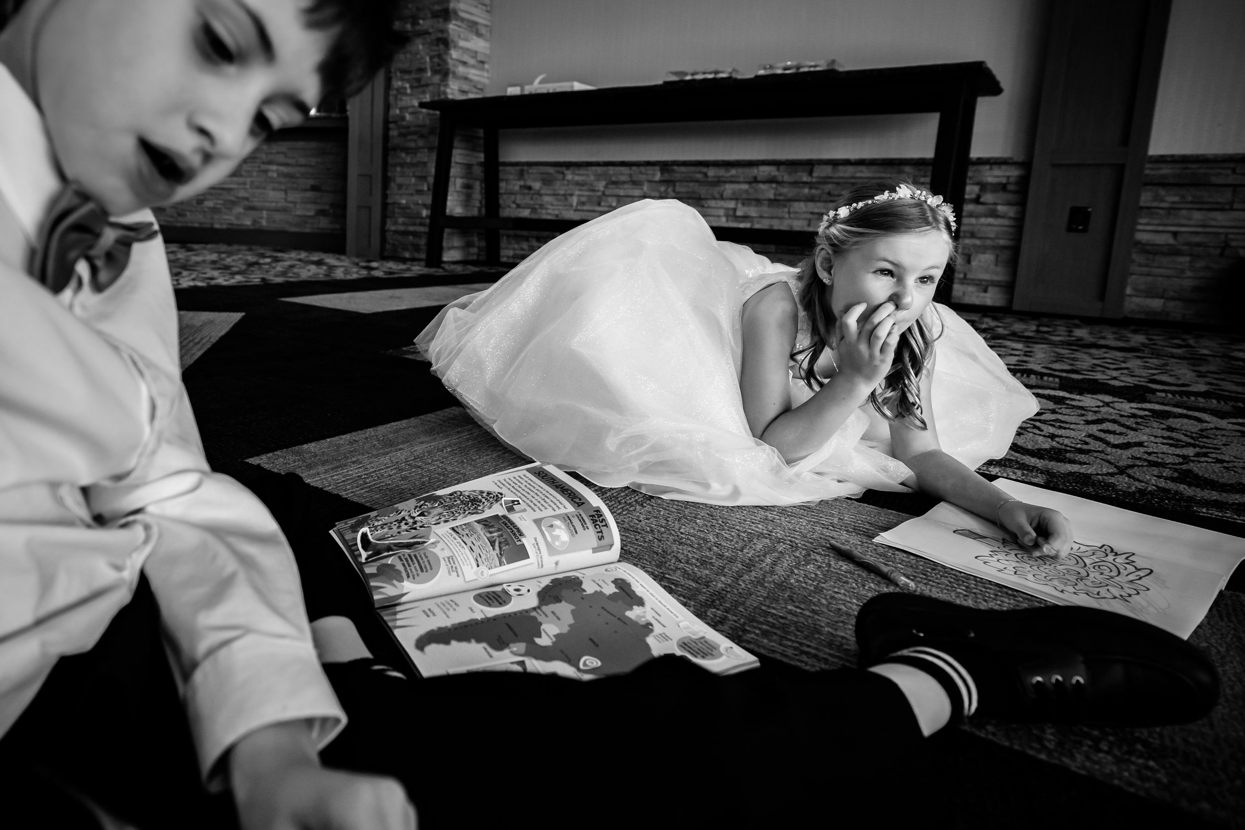 Bride lying on floor in white gown coloring with young boy at wedding reception in black and white.