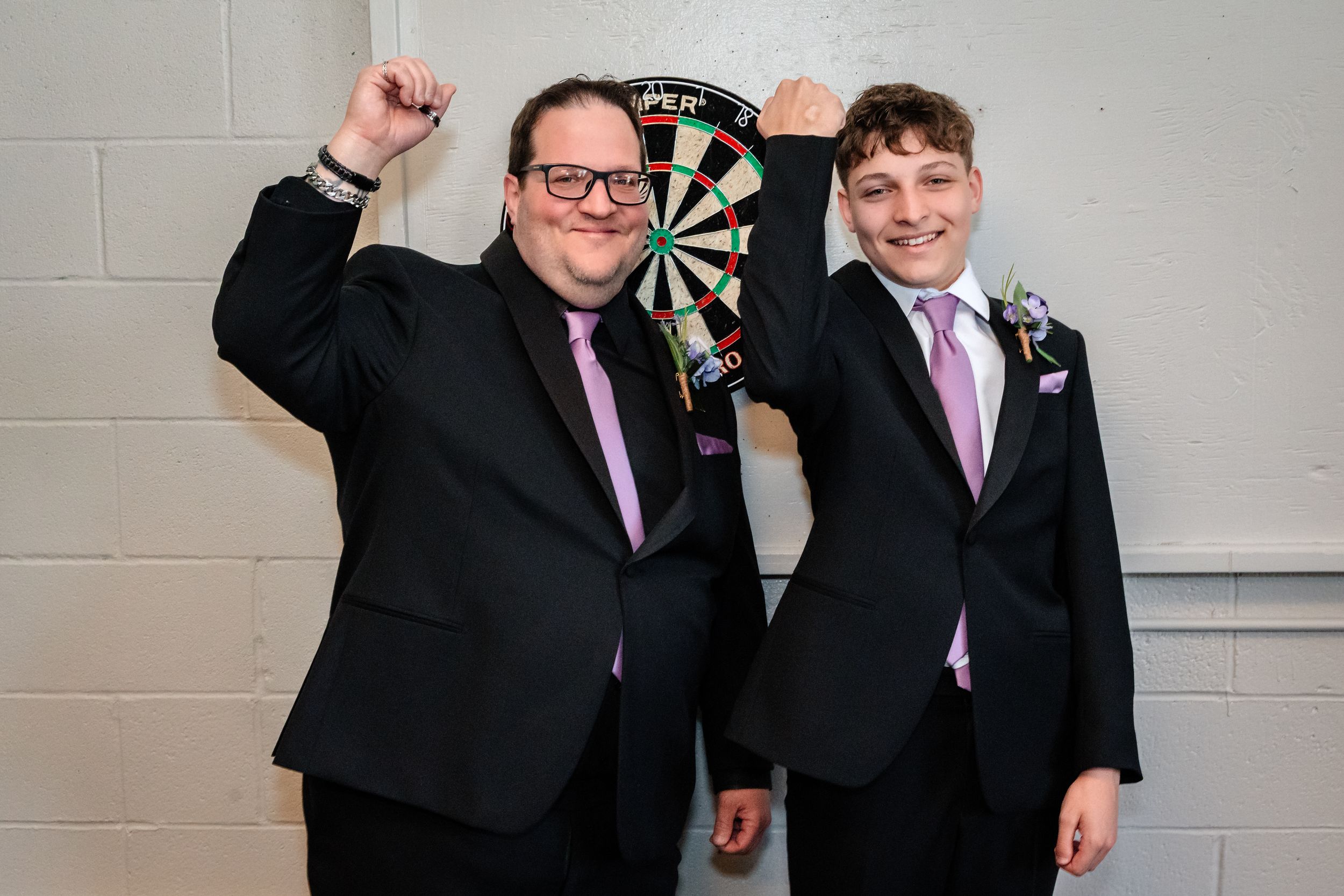Two groomsmen in black suits with purple ties celebrating in front of dartboard at wedding reception.