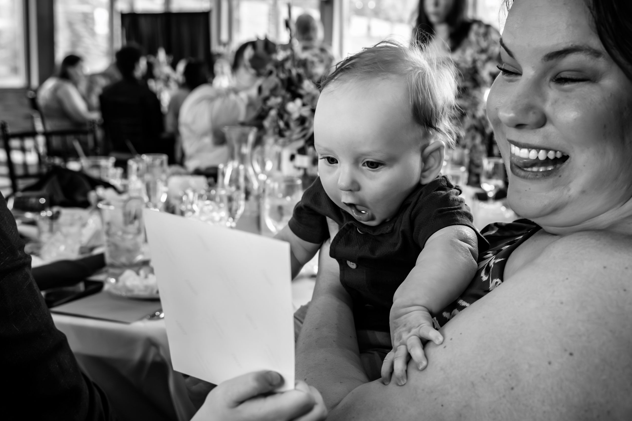 Black and white photo of a laughing woman holding a curious baby at a wedding reception.