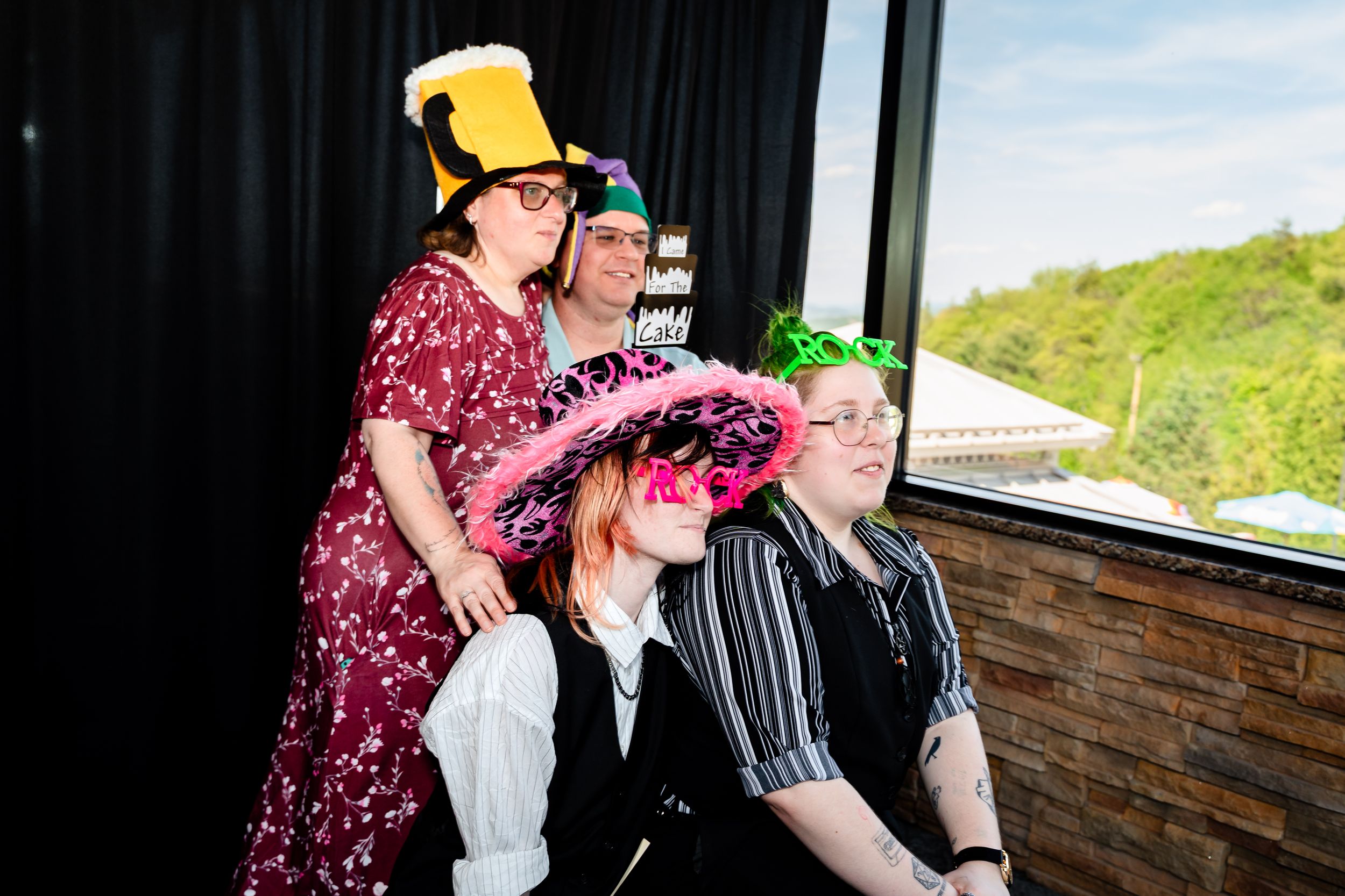 Four wedding guests laugh wearing colorful novelty hats at photo booth during reception near window.