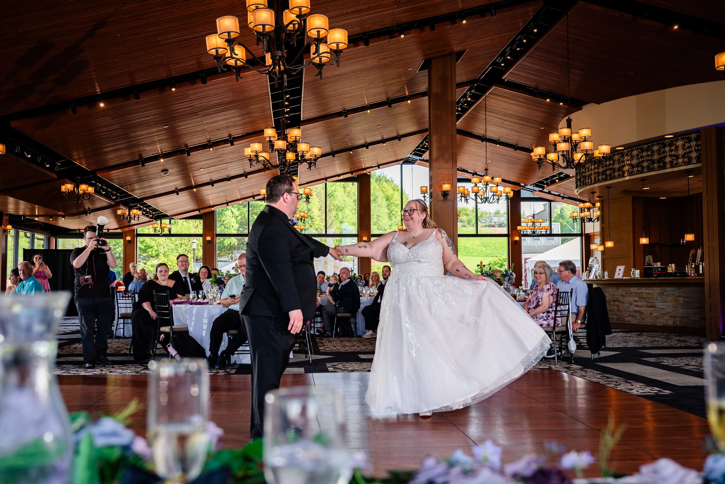 Bride and groom share their first dance in an elegant wood-beamed reception hall with chandelier lighting.