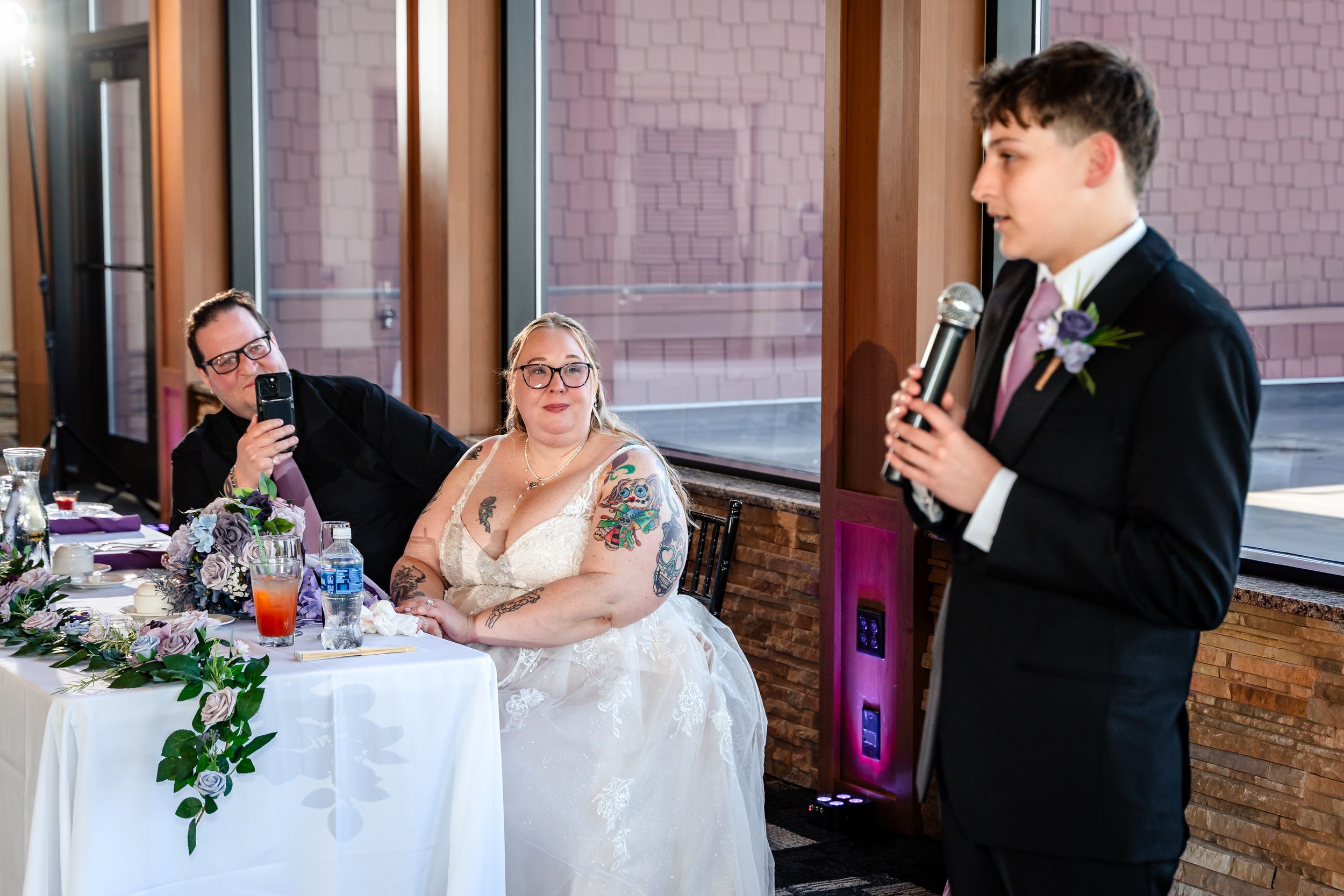 Young man gives speech at microphone while bride and groom listen at decorated wedding head table.