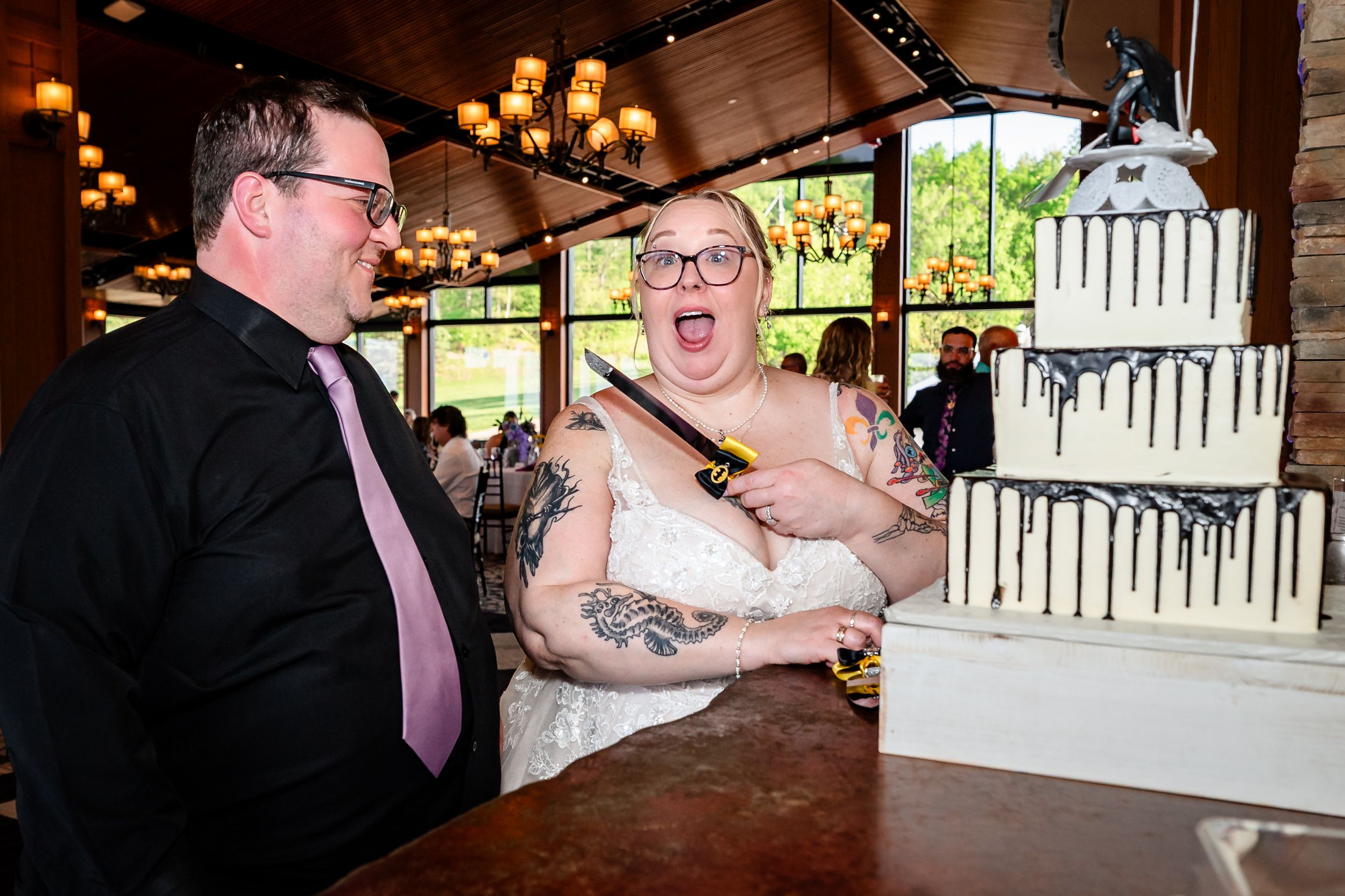 Excited bride with tattoos and groom cut white drip wedding cake with Batman topper at reception.